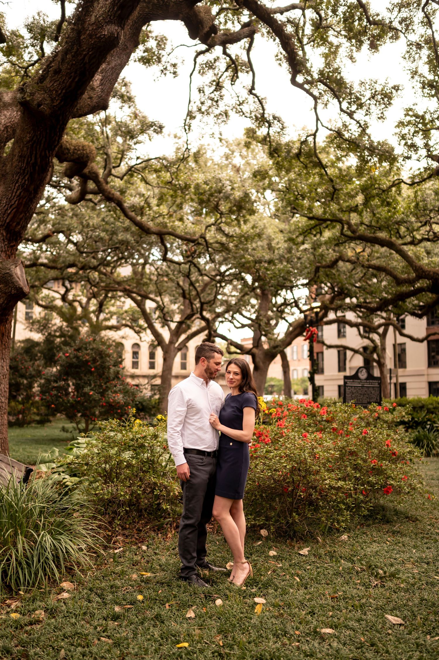 A couple stands together under sprawling oak trees in a garden setting with red flowers and historic architecture nearby.
