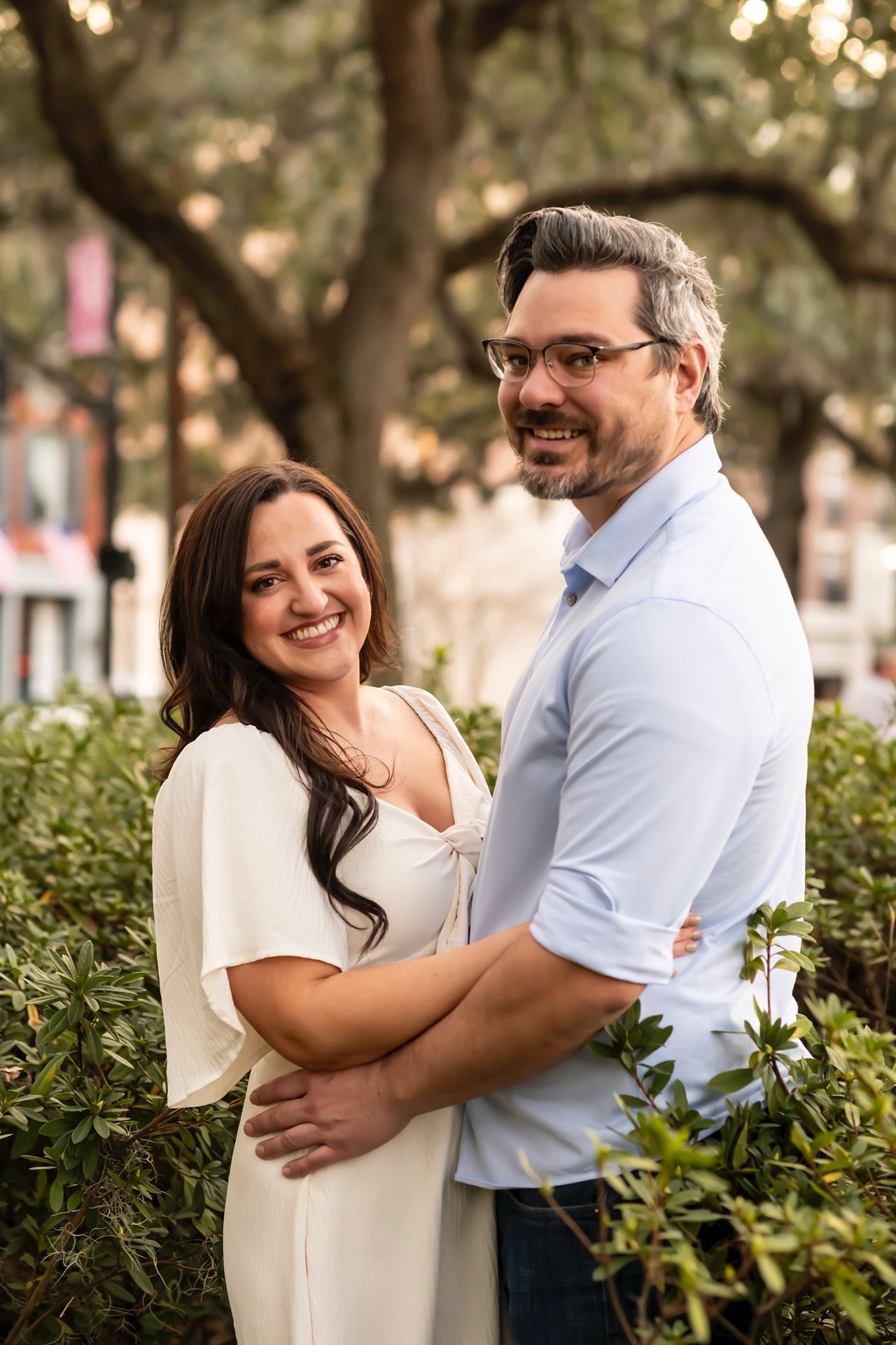 Couple embraces outdoors in casual attire while standing in front of lush greenery during sunset.
