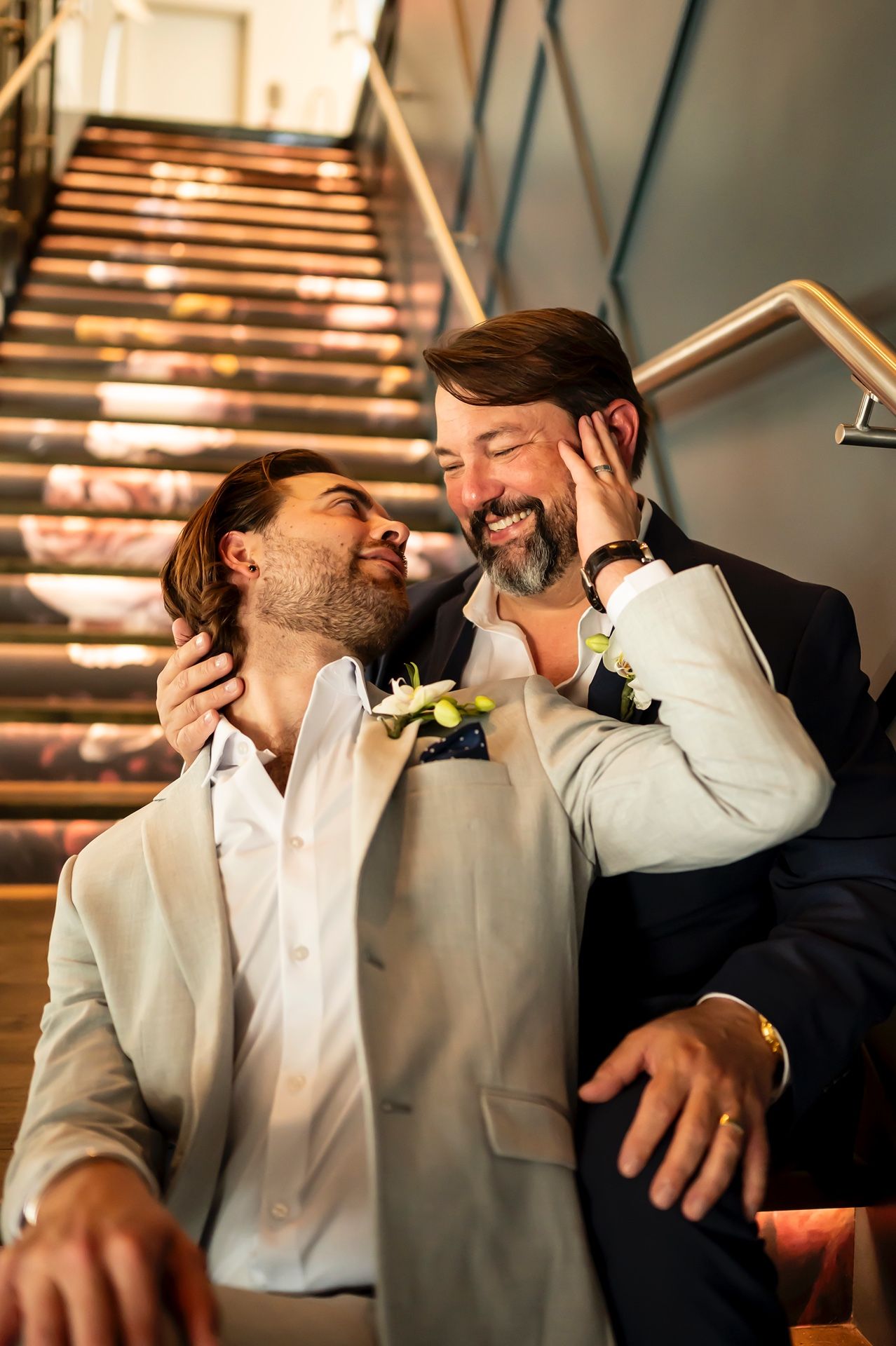Two people in suits sharing an intimate moment on a staircase with warm lighting and boutonnieres.