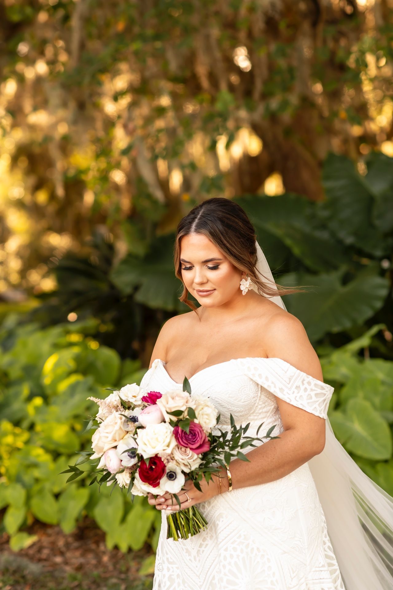 A bride in an off-shoulder lace wedding dress holds a romantic bouquet of white and burgundy flowers in a garden setting.