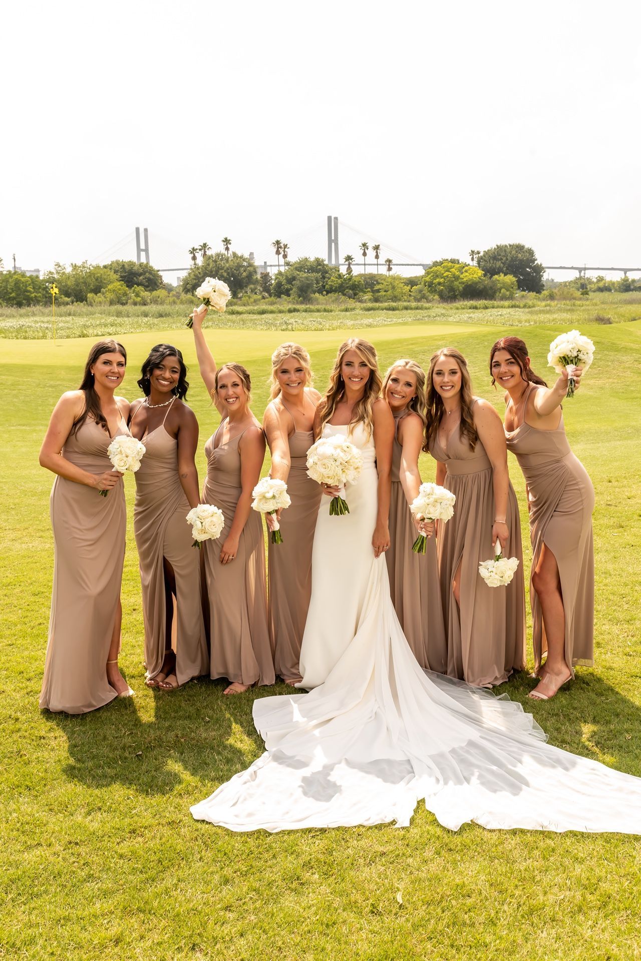 A bridal party in taupe bridesmaid dresses and a white wedding gown pose with bouquets on a sunny golf course.