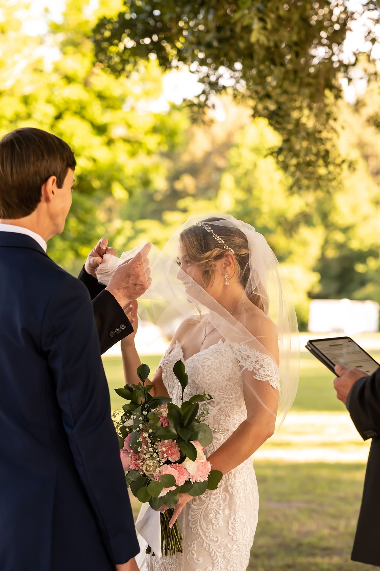 A couple exchanges wedding vows during an outdoor ceremony in golden sunlight as an officiant holds a tablet.