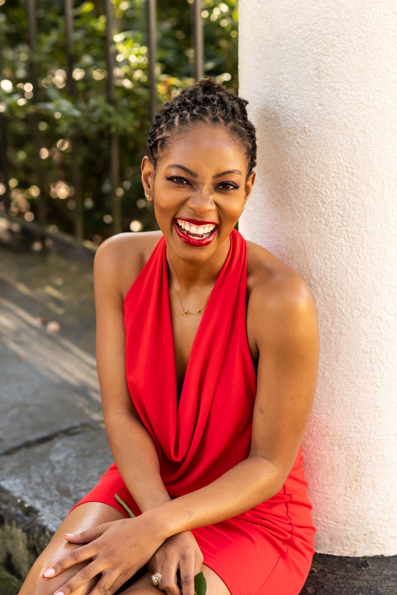 A joyful expression radiates as someone in a red halter dress sits by a white column outdoors.
