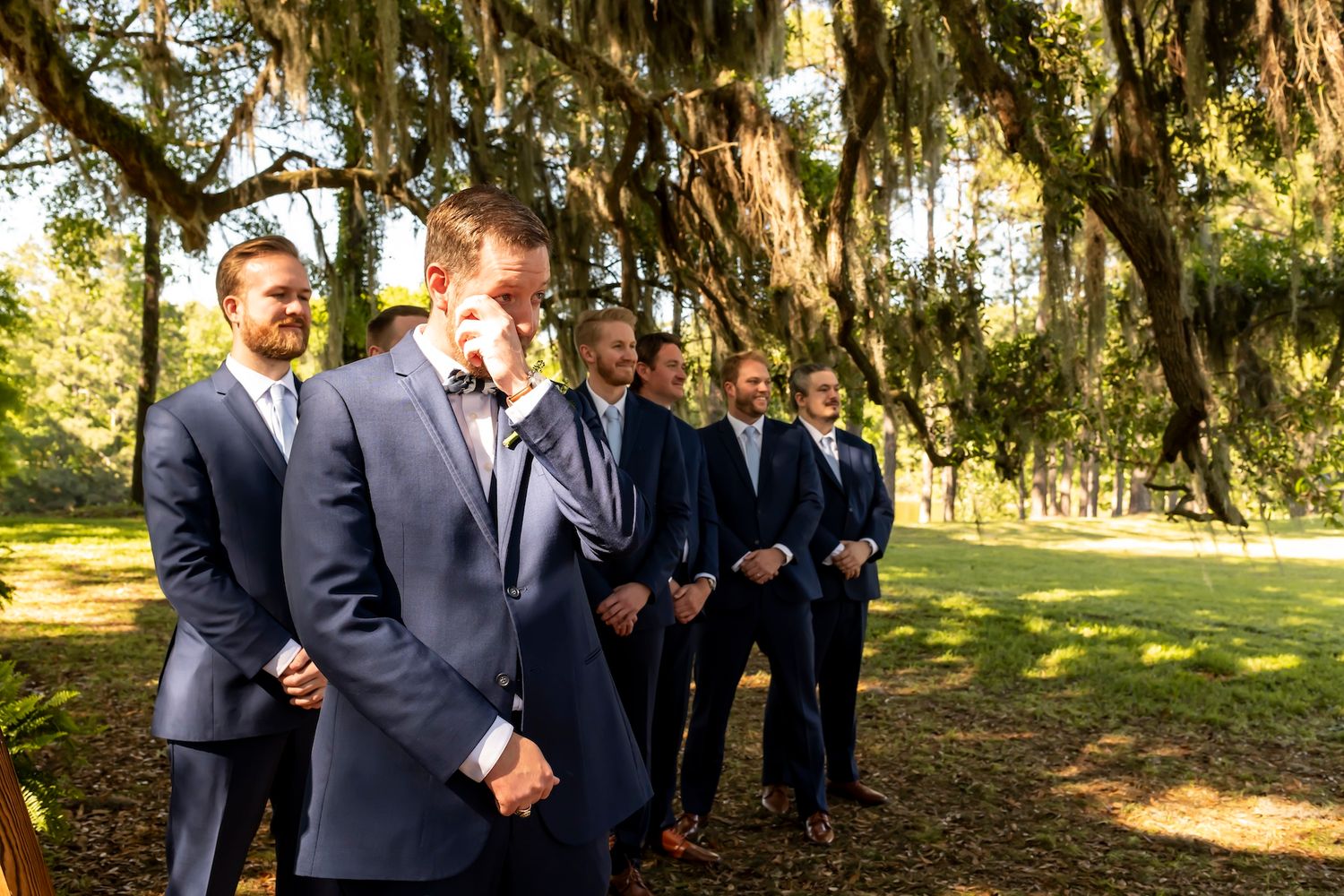 Groomsmen standing in line on sunny day under trees draped with Spanish moss in outdoor garden setting.