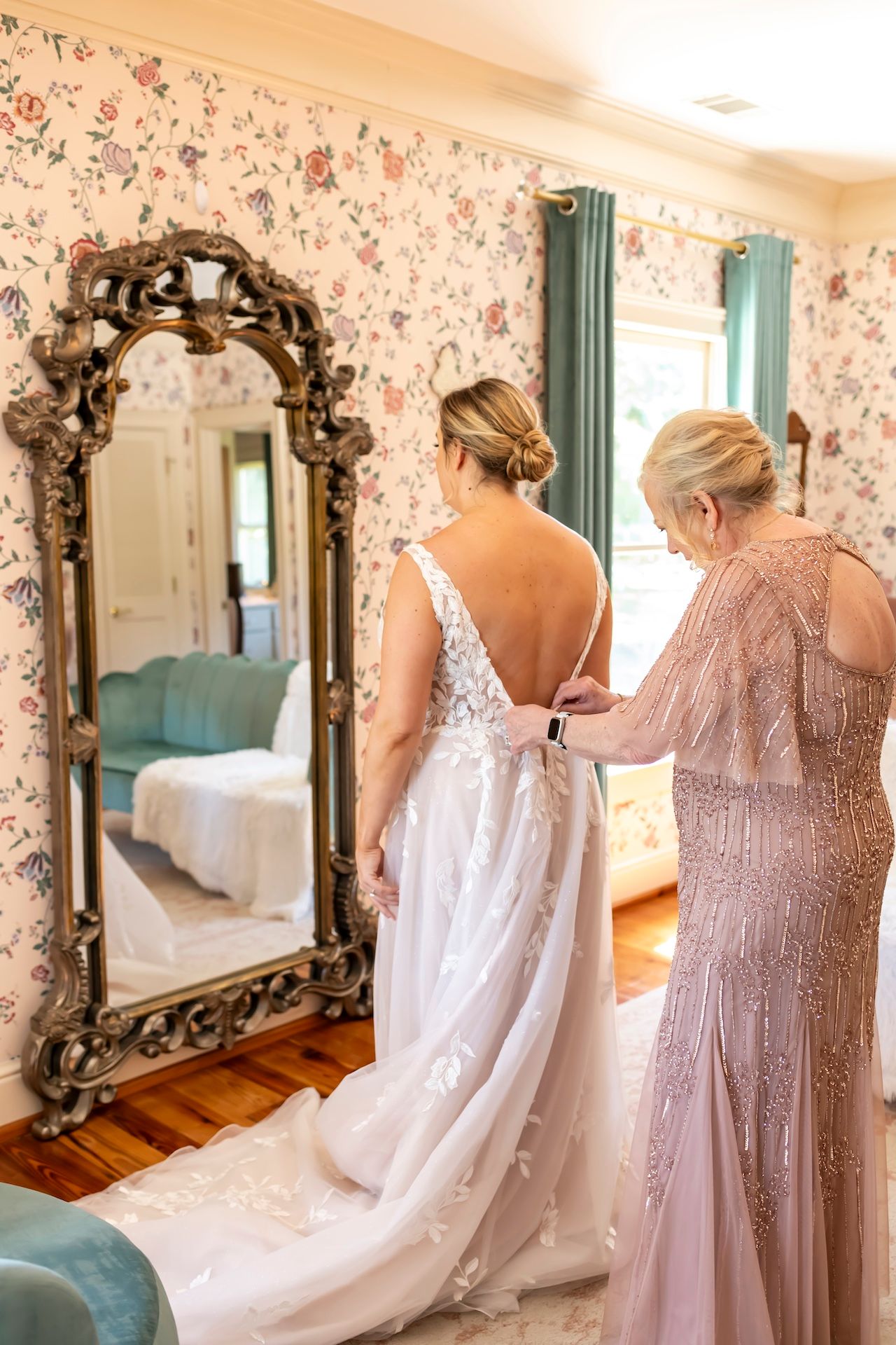 A woman in a white wedding dress stands before an ornate mirror while someone in pink beaded gown helps with the dress.