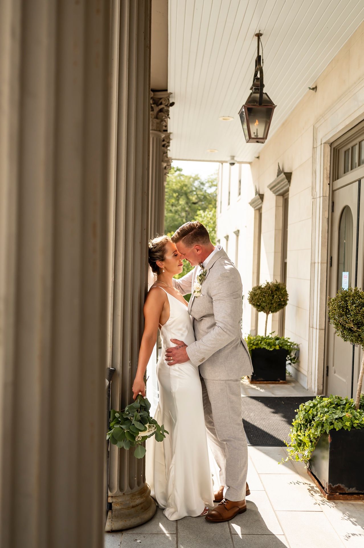 Newlyweds share an intimate moment on an elegant porch with white columns and hanging lanterns.