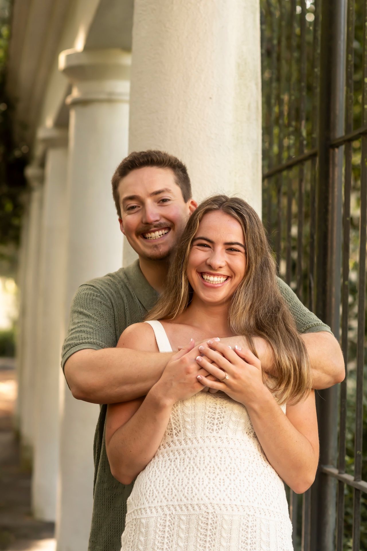 A happy couple embraces outdoors near white columns, with her wearing a white dress and him in an olive green shirt.