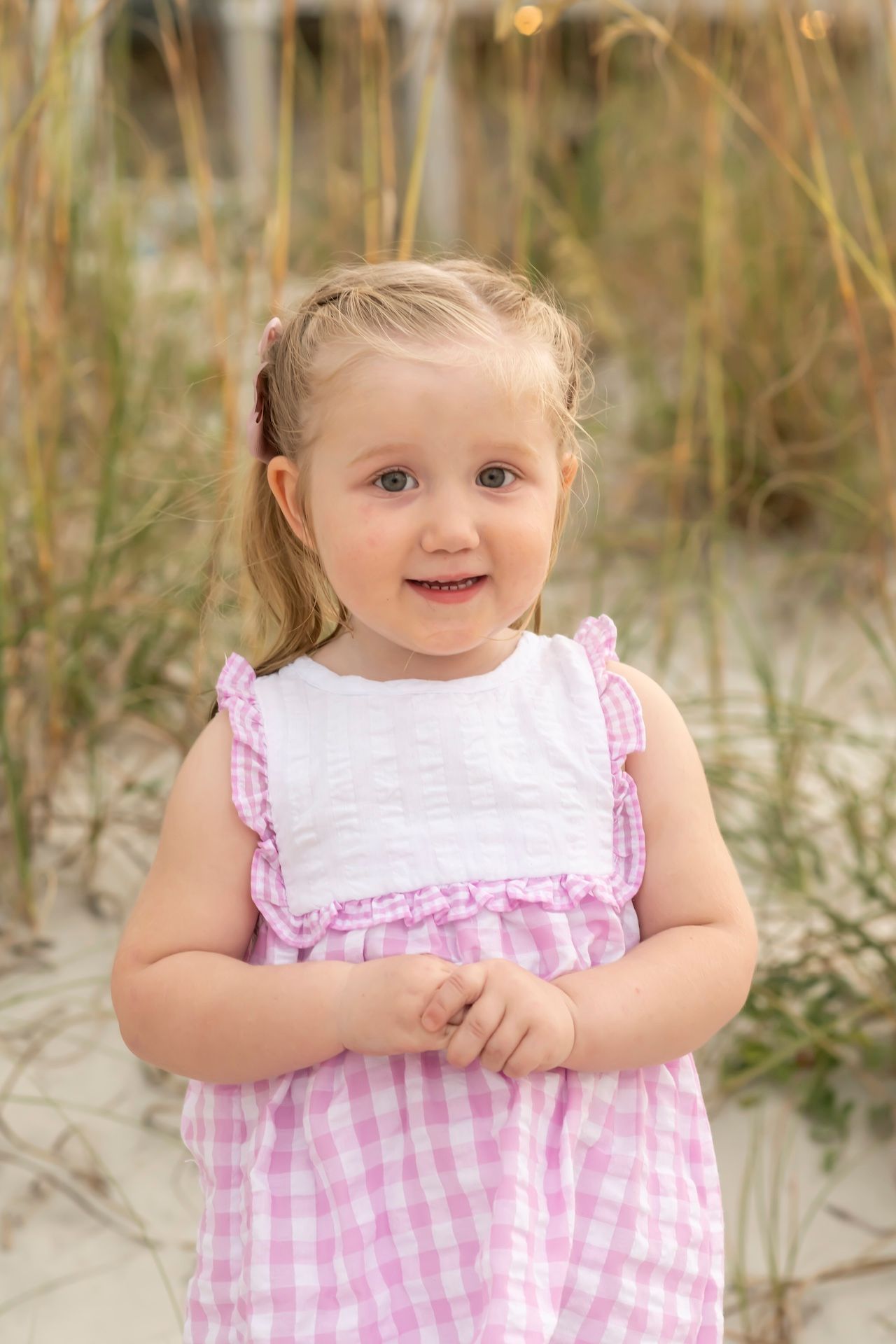 A child in a pink gingham dress smiles while standing among beach grass and sand dunes.
