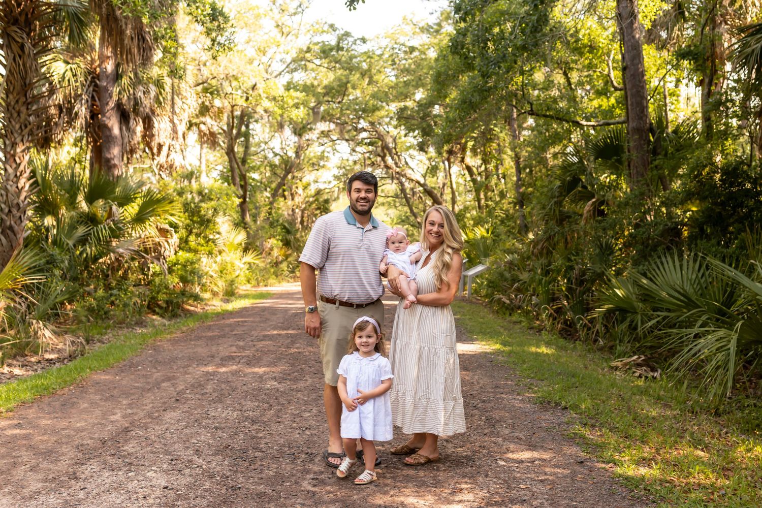 Family in white outfits walking and posing together on a sunlit nature trail surrounded by tropical greenery.