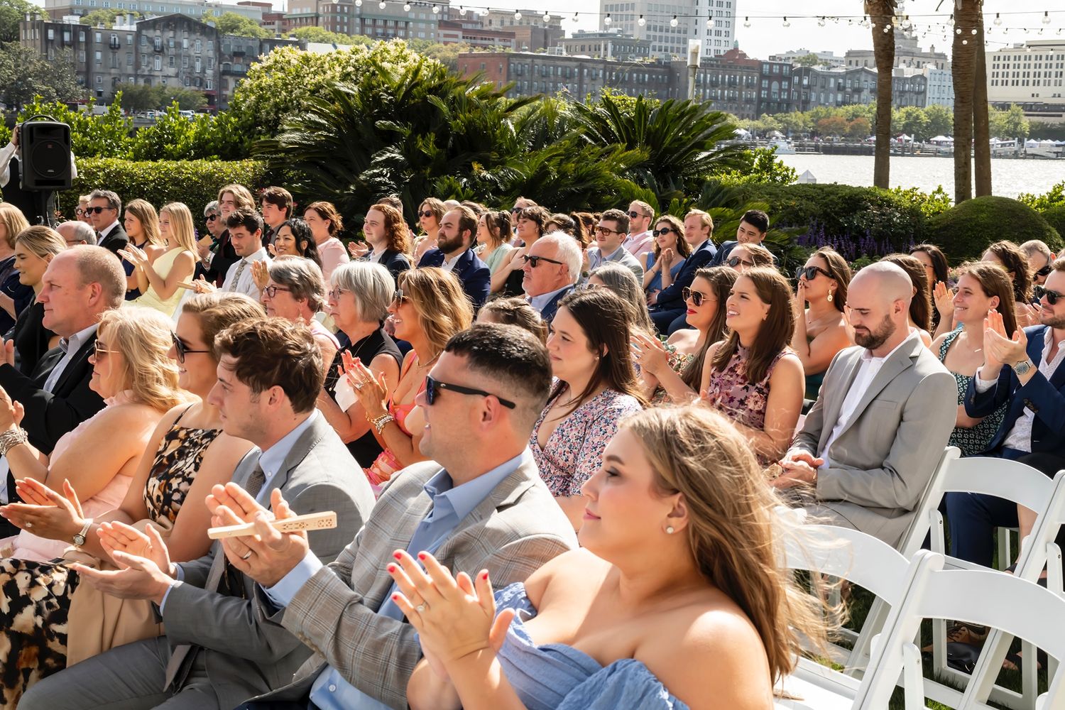 Outdoor wedding guests sitting in white chairs applaud during a sunny ceremony with tropical plants in the background.