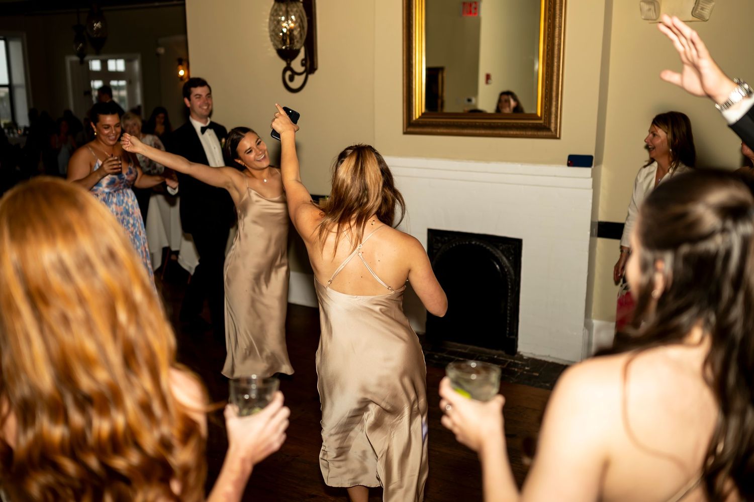 Party guests with drinks dance and celebrate at an elegant indoor reception with a fireplace and framed artwork.