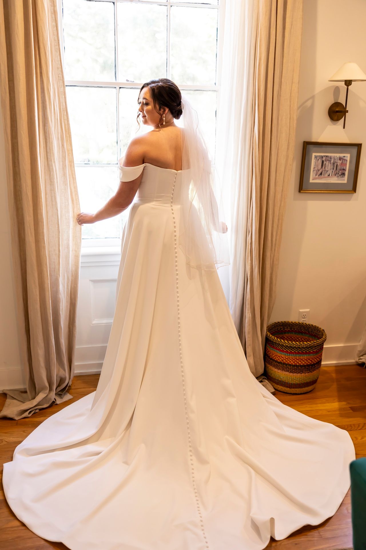 An elegant off-shoulder wedding gown with button details and flowing train is shown from behind near a sunny window.