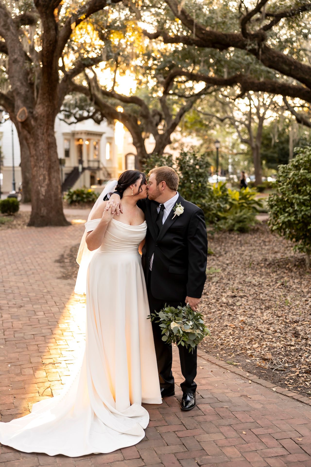 A romantic sunset wedding photo showing a couple embracing on a brick path under majestic oak trees.