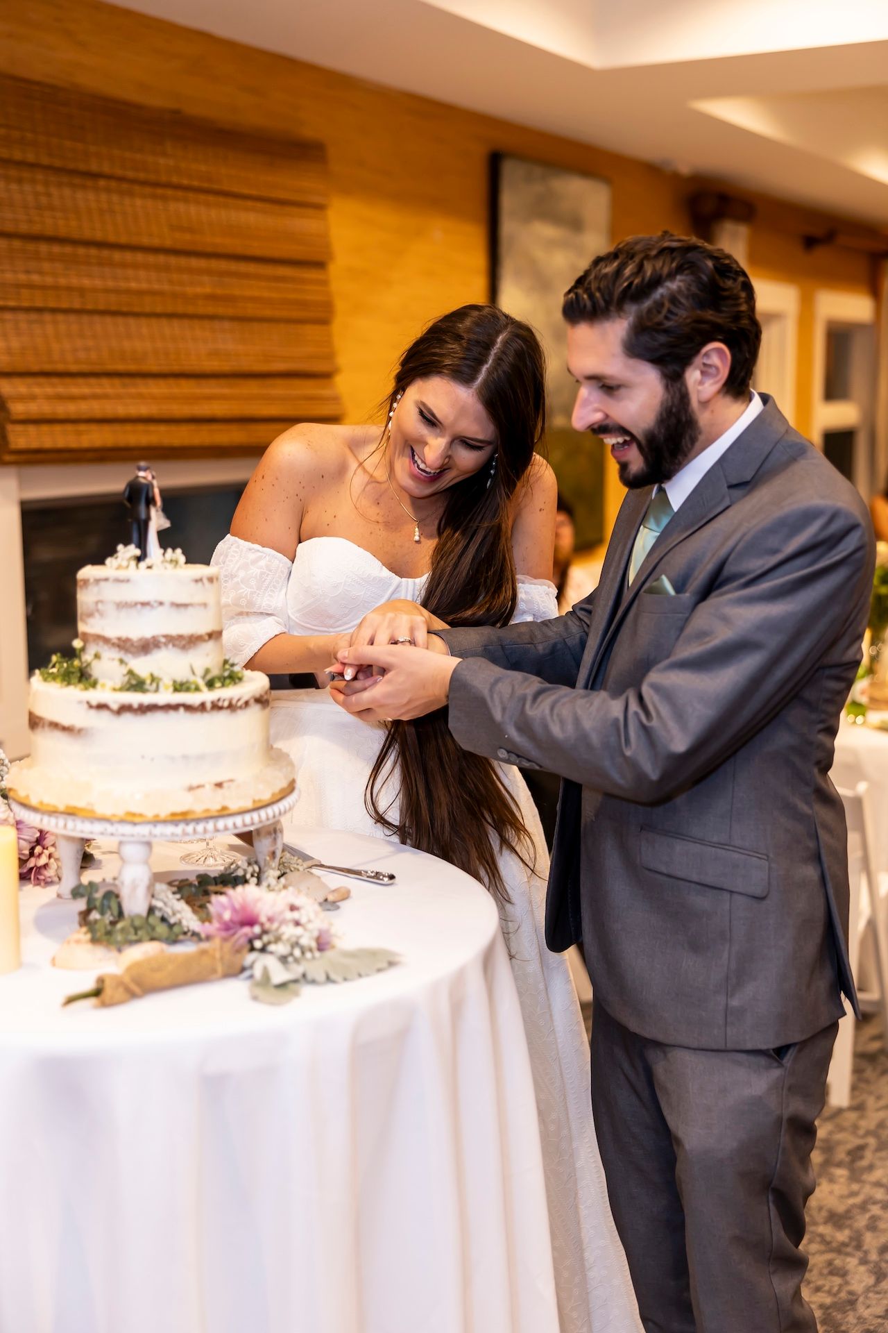 Newlyweds cutting their white tiered wedding cake together at an indoor reception.