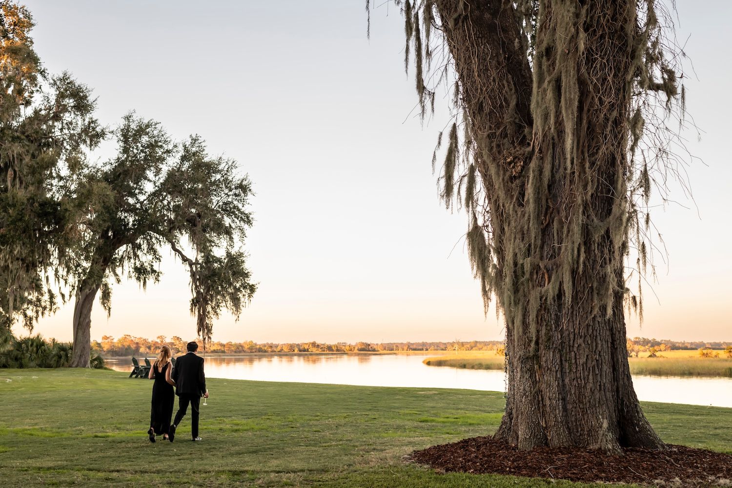 A couple stands by a lake at sunset with spanish moss hanging from a large oak tree in the foreground.