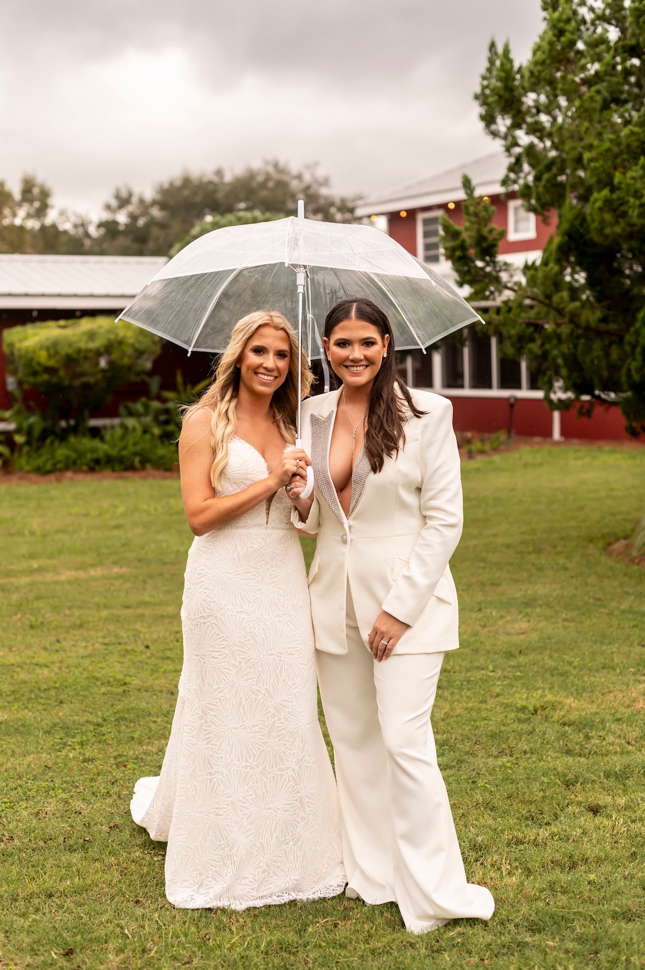 Two people in white wedding attire share a clear umbrella while posing on a green lawn with a building in the background.