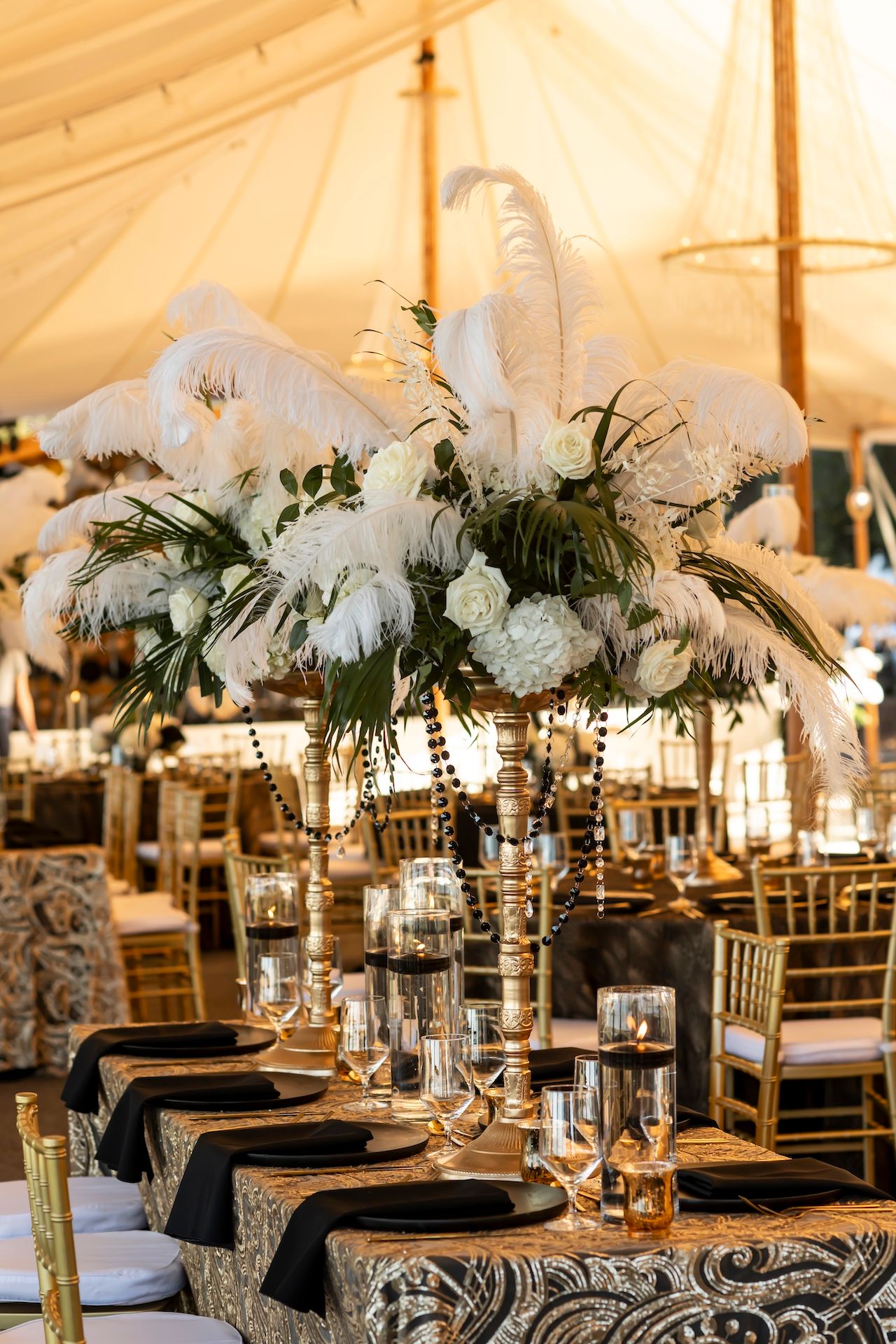 Elegant wedding table setting with tall white feather centerpiece and crystal decor under a white tent.