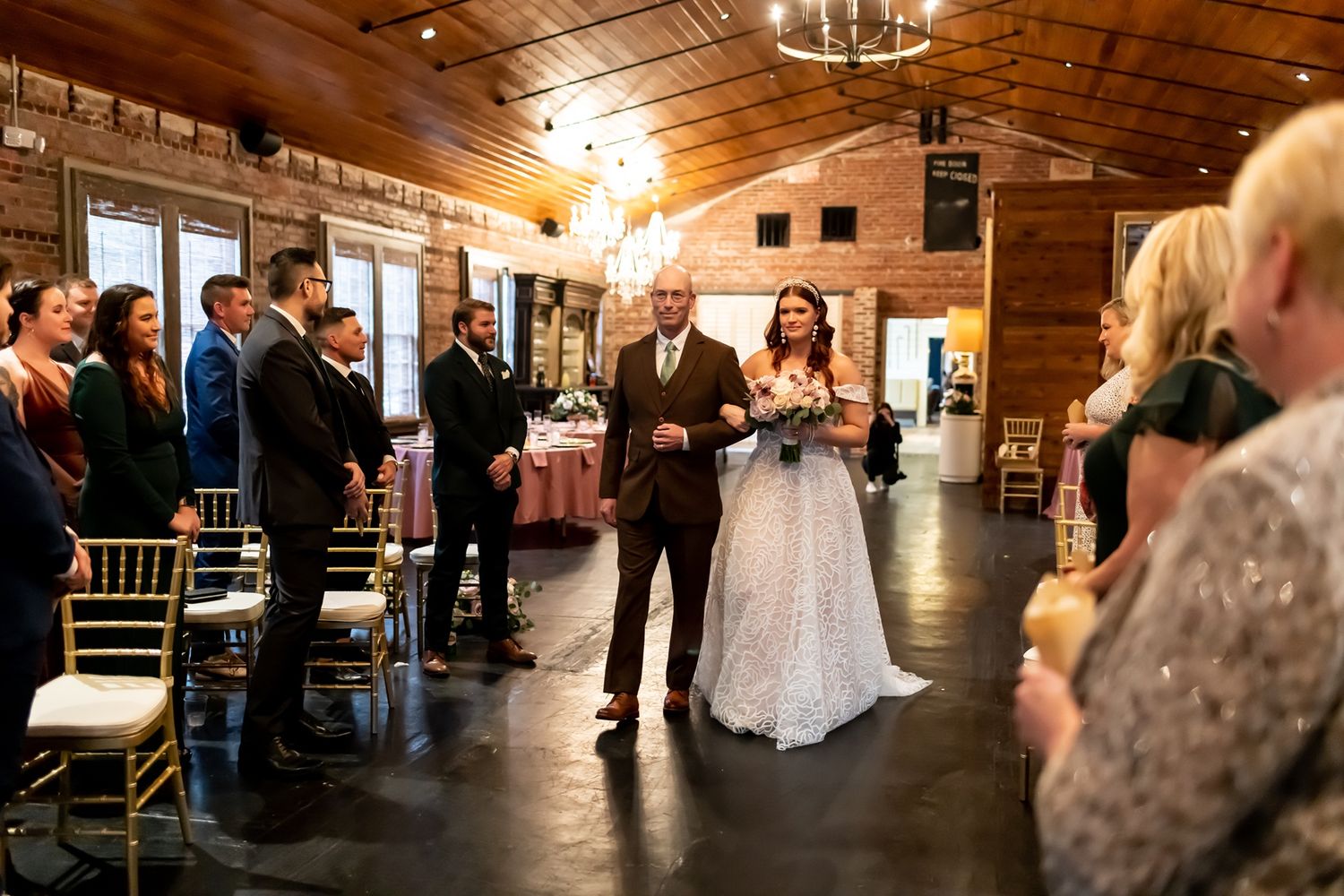Wedding ceremony processional in rustic wooden barn venue with exposed beam ceiling and elegant lighting fixtures.
