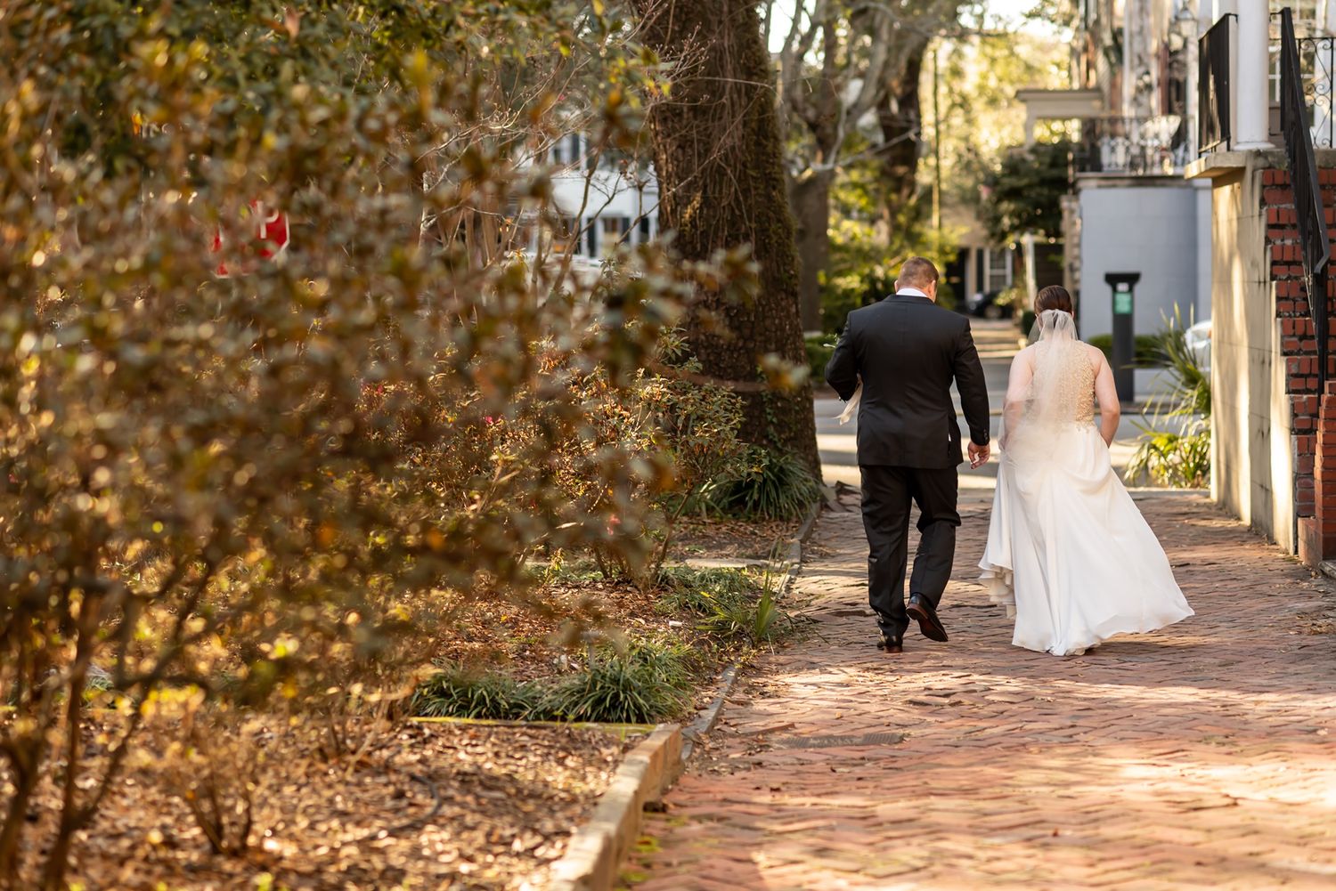 Couple in formal attire walking down a brick pathway lined with autumn foliage and historic buildings.