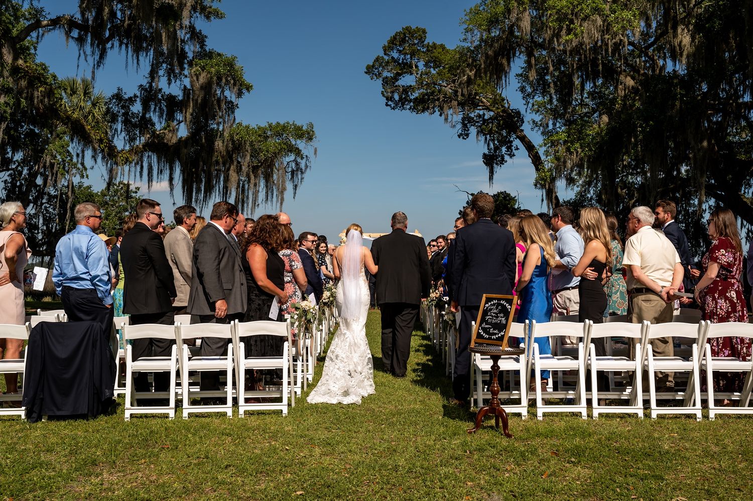 An outdoor wedding ceremony with white chairs set up on grass under moss-draped oak trees in a Southern garden setting.