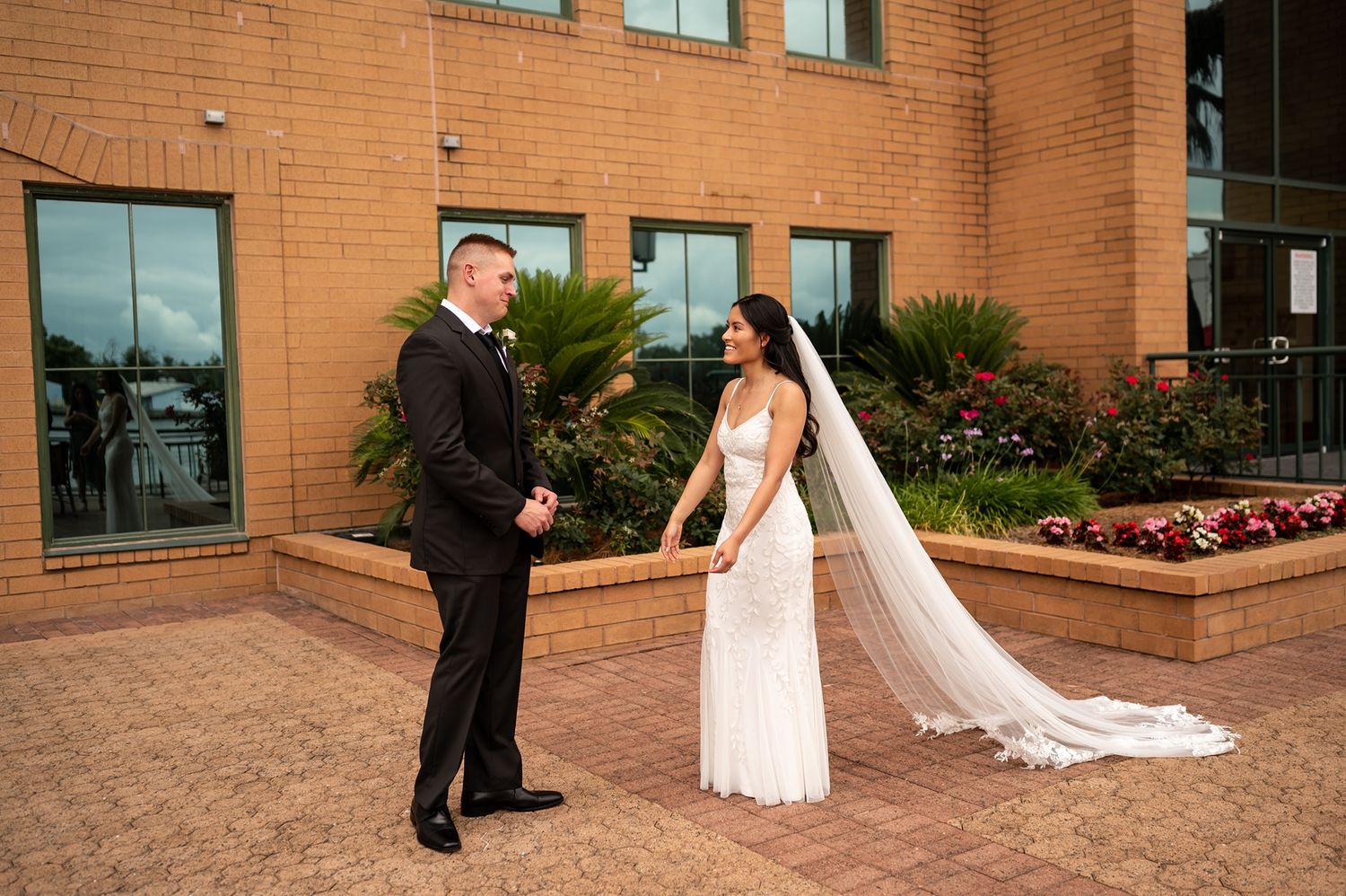 A couple shares a joyful moment in their wedding attire outside a brick building with landscaped gardens.