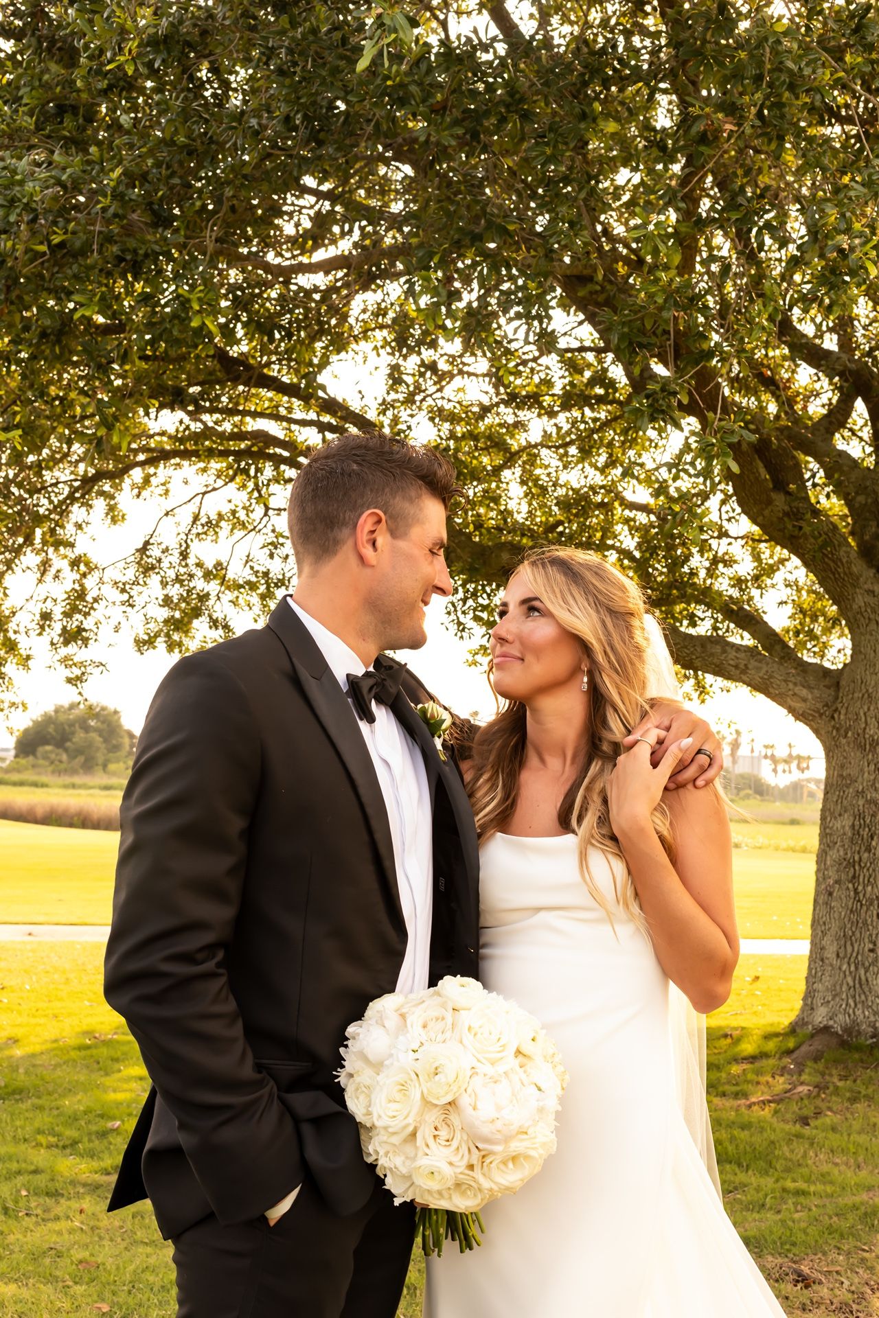 A couple share an intimate moment at sunset under a large oak tree on their wedding day.