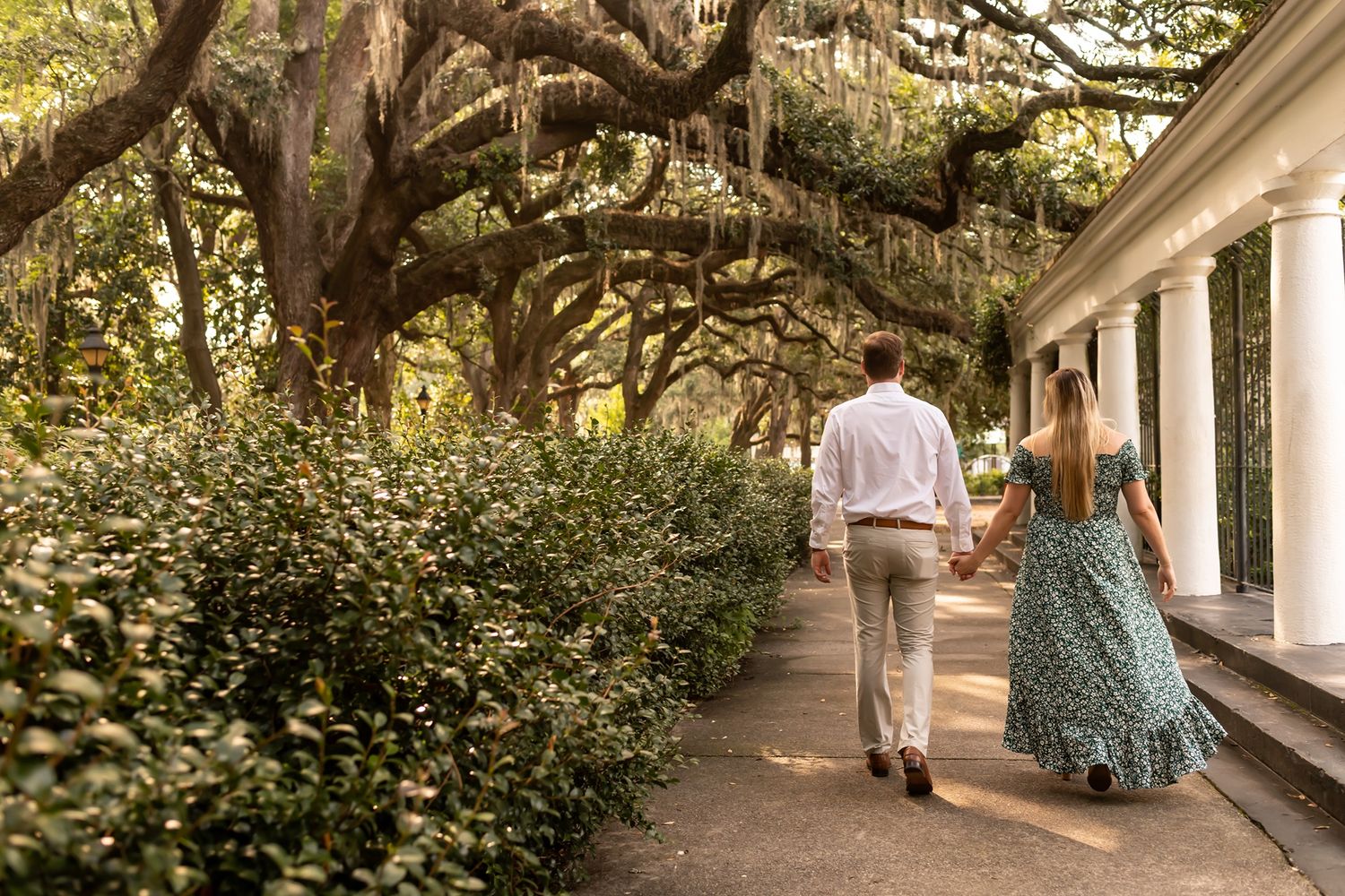 A couple strolls down a scenic garden path lined with oak trees draped in Spanish moss beside a white columned building.