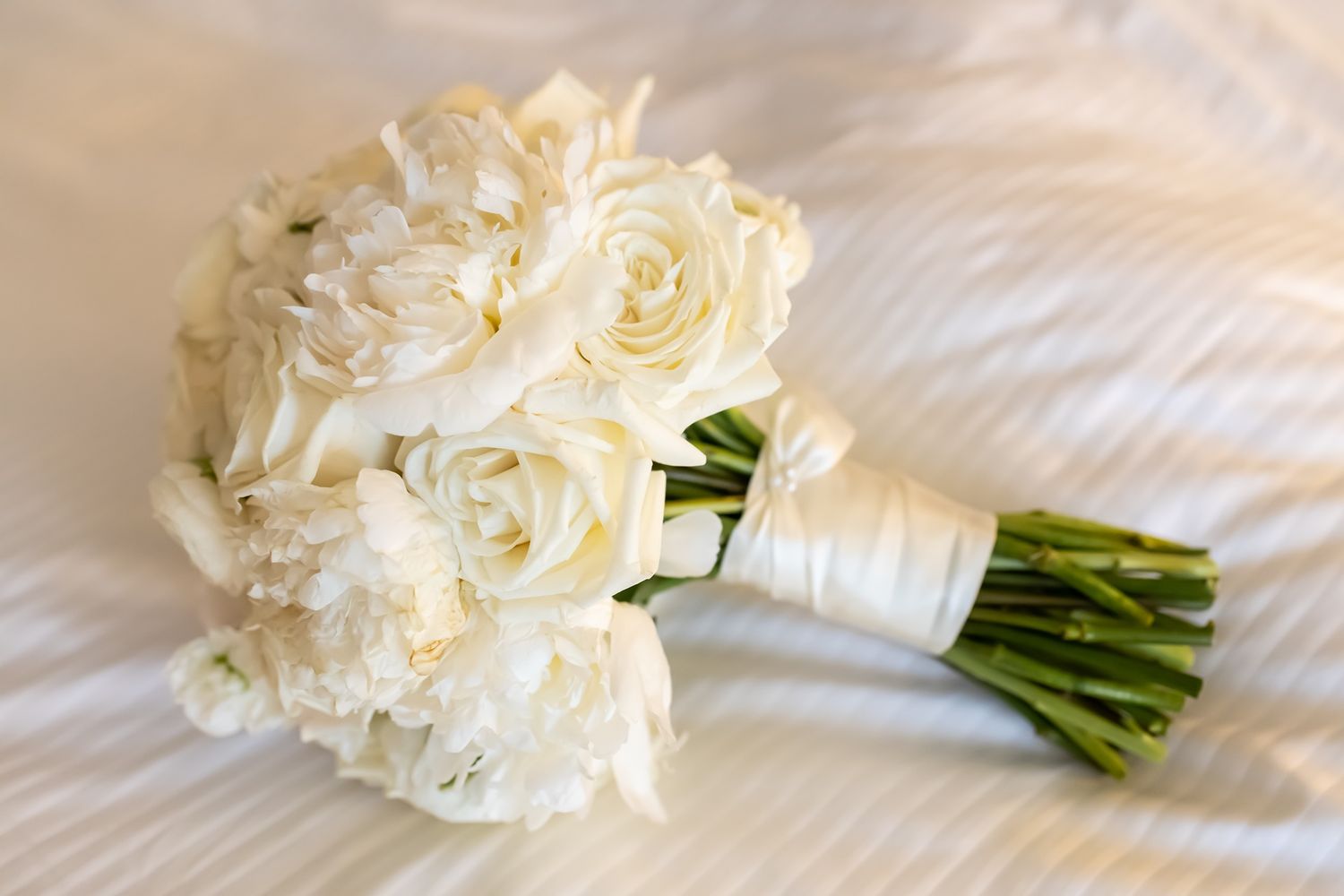 Elegant white rose bridal bouquet wrapped in satin ribbon laying on white textured fabric.