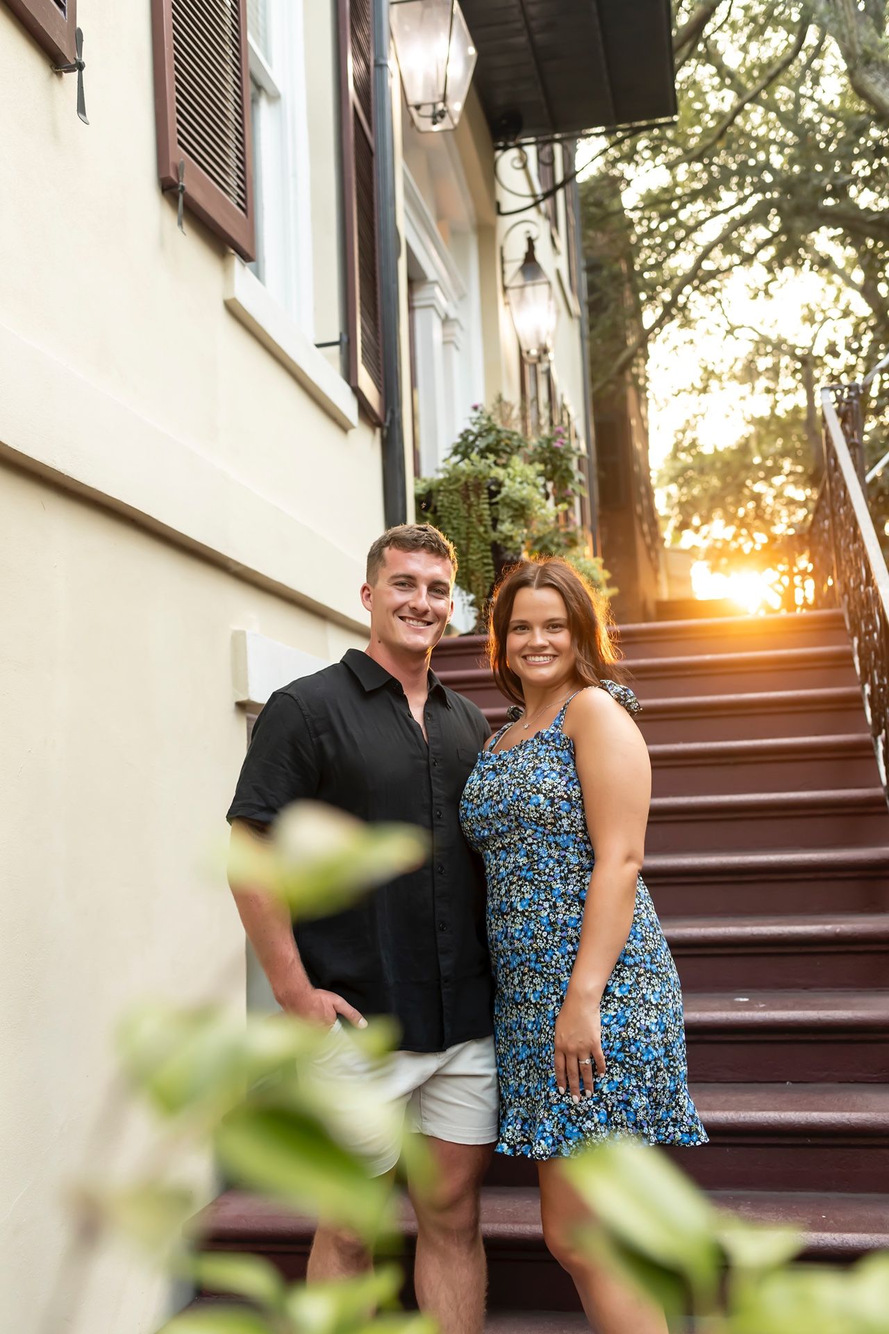A couple stands together on outdoor steps by a white building as golden sunlight filters through trees at sunset.