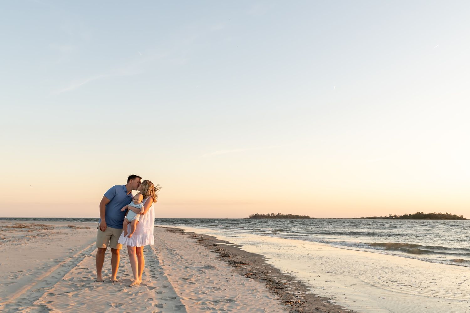 A romantic couple stands silhouetted against a serene sunset on a sandy beach during golden hour.