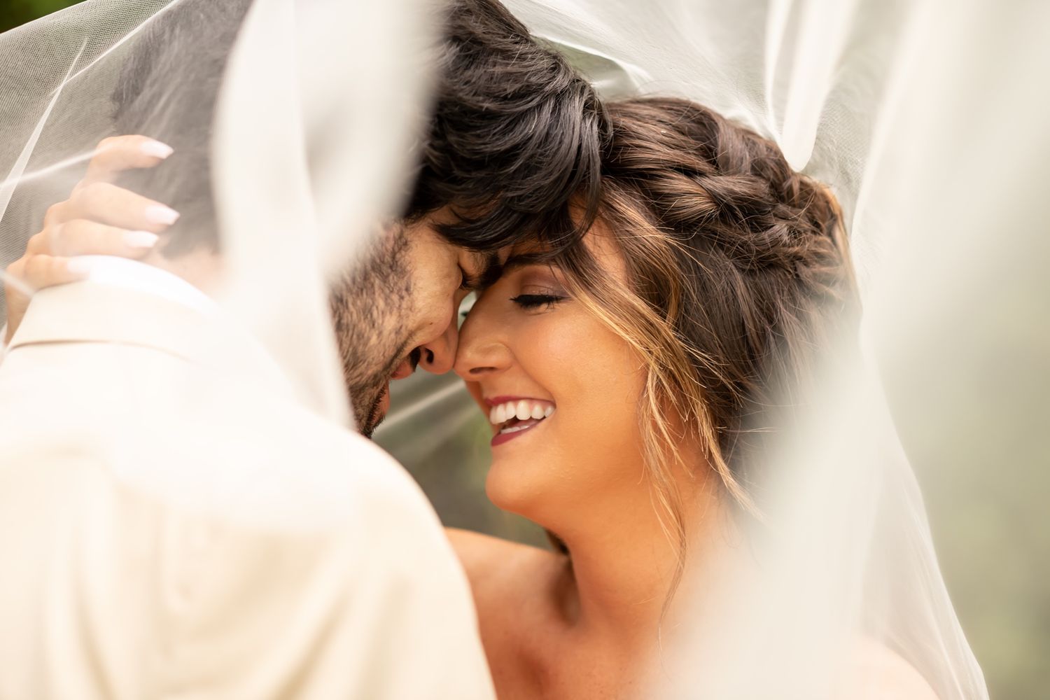 Close up romantic moment under wedding veil with bride smiling in warm sunlight.