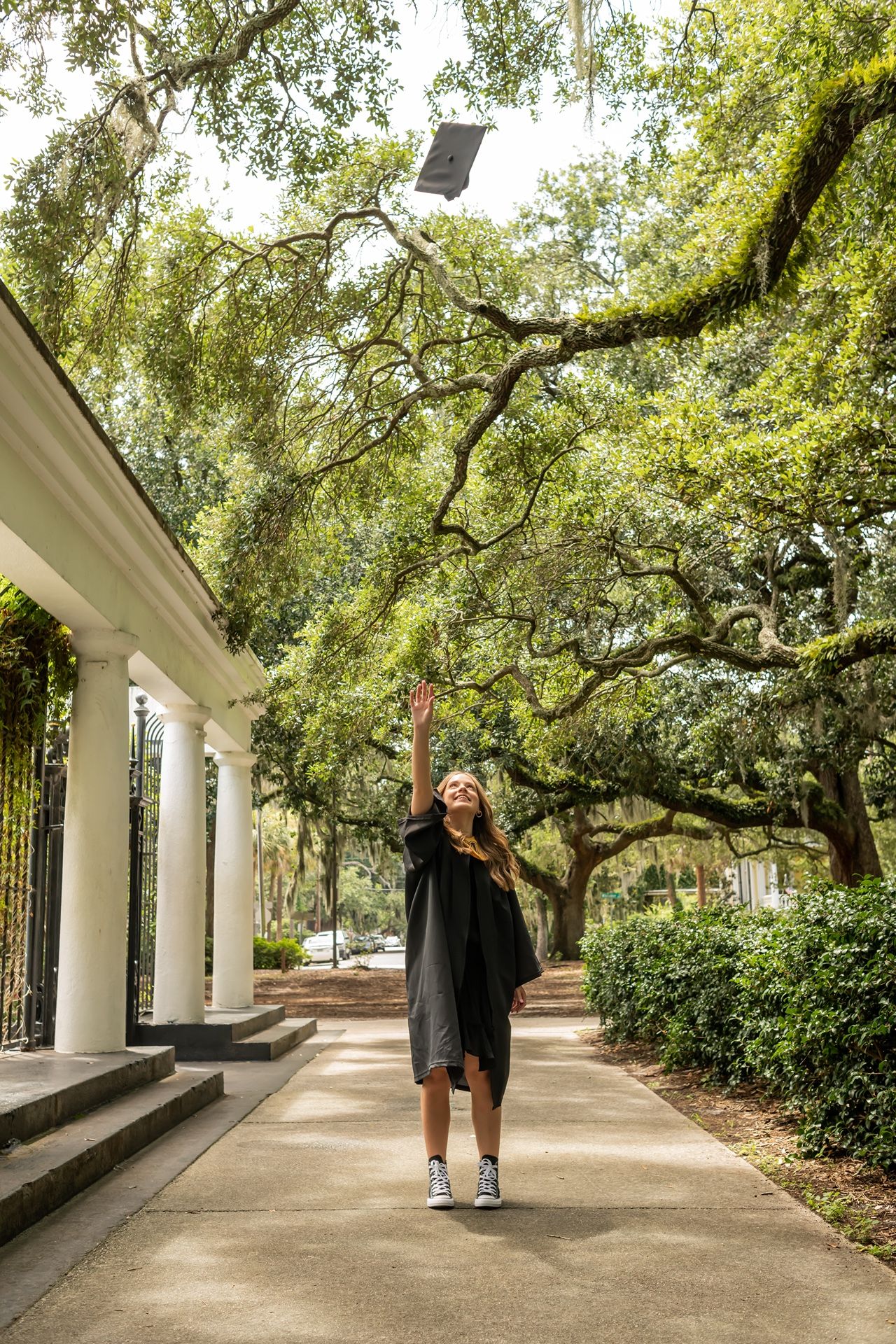 Graduate in black cap and gown celebrating on tree-lined campus walkway with historic columns and Spanish moss overhead.
