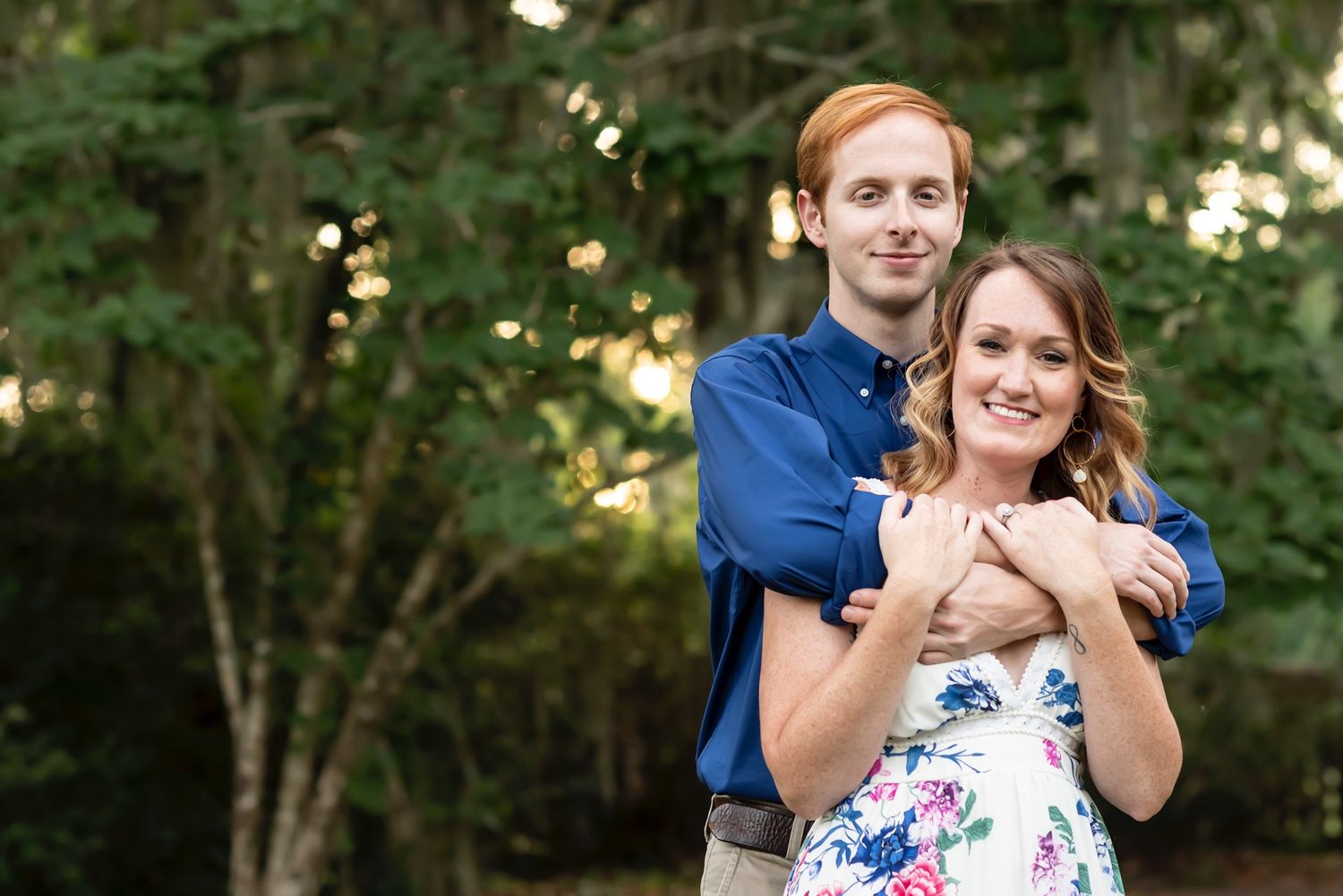A couple embraces outdoors in a natural setting with trees and soft lighting in the background.