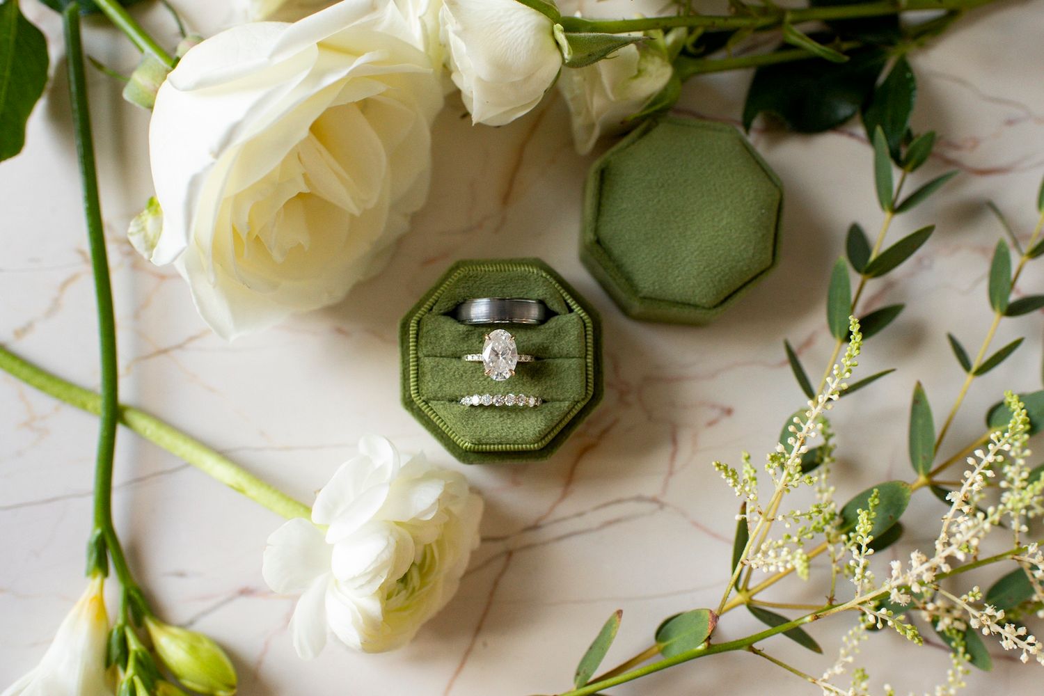 Elegant flat lay of white roses, green leaves, and a hexagonal ring box on a white marble surface.