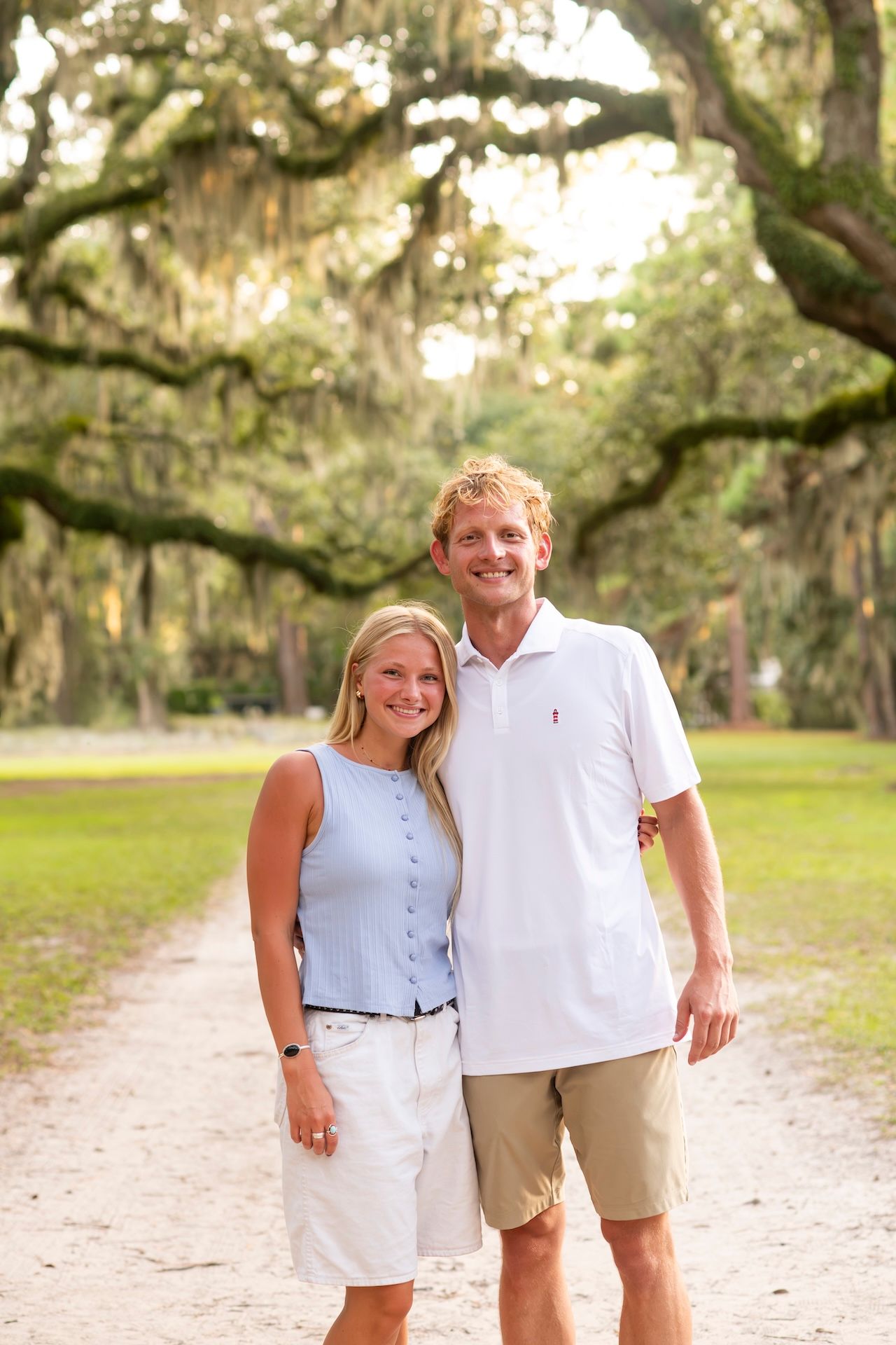 Smiling couple posing on a scenic tree-lined path with Spanish moss-draped oak trees in a park.
