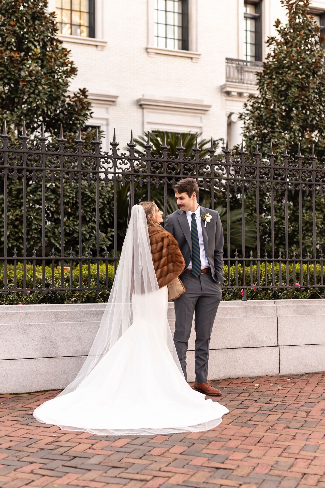 A romantic wedding couple stands together on a brick patio beside a decorative iron fence and white building.