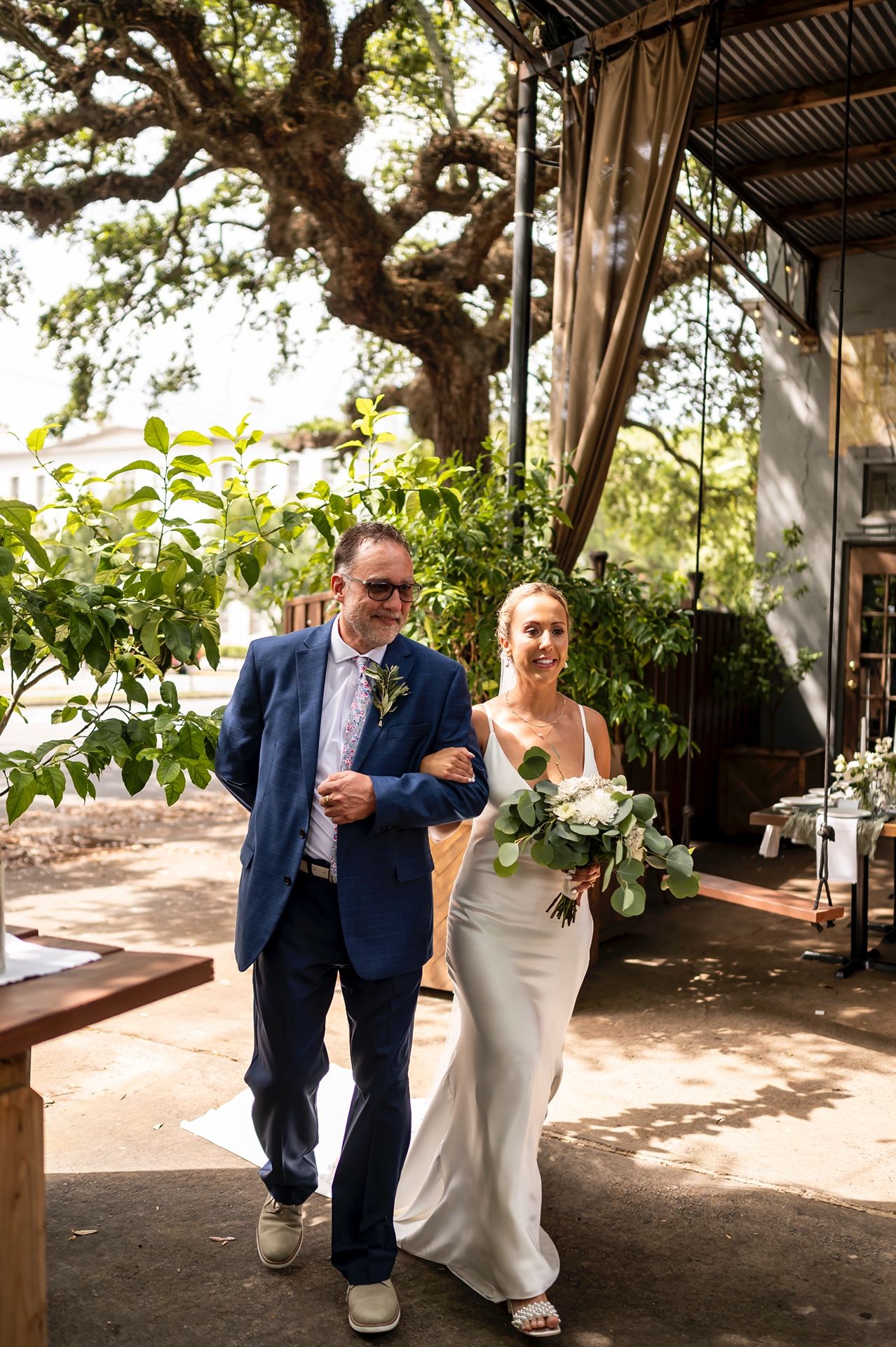 A pair walks down a shaded garden path under a large oak tree at an outdoor wedding venue.