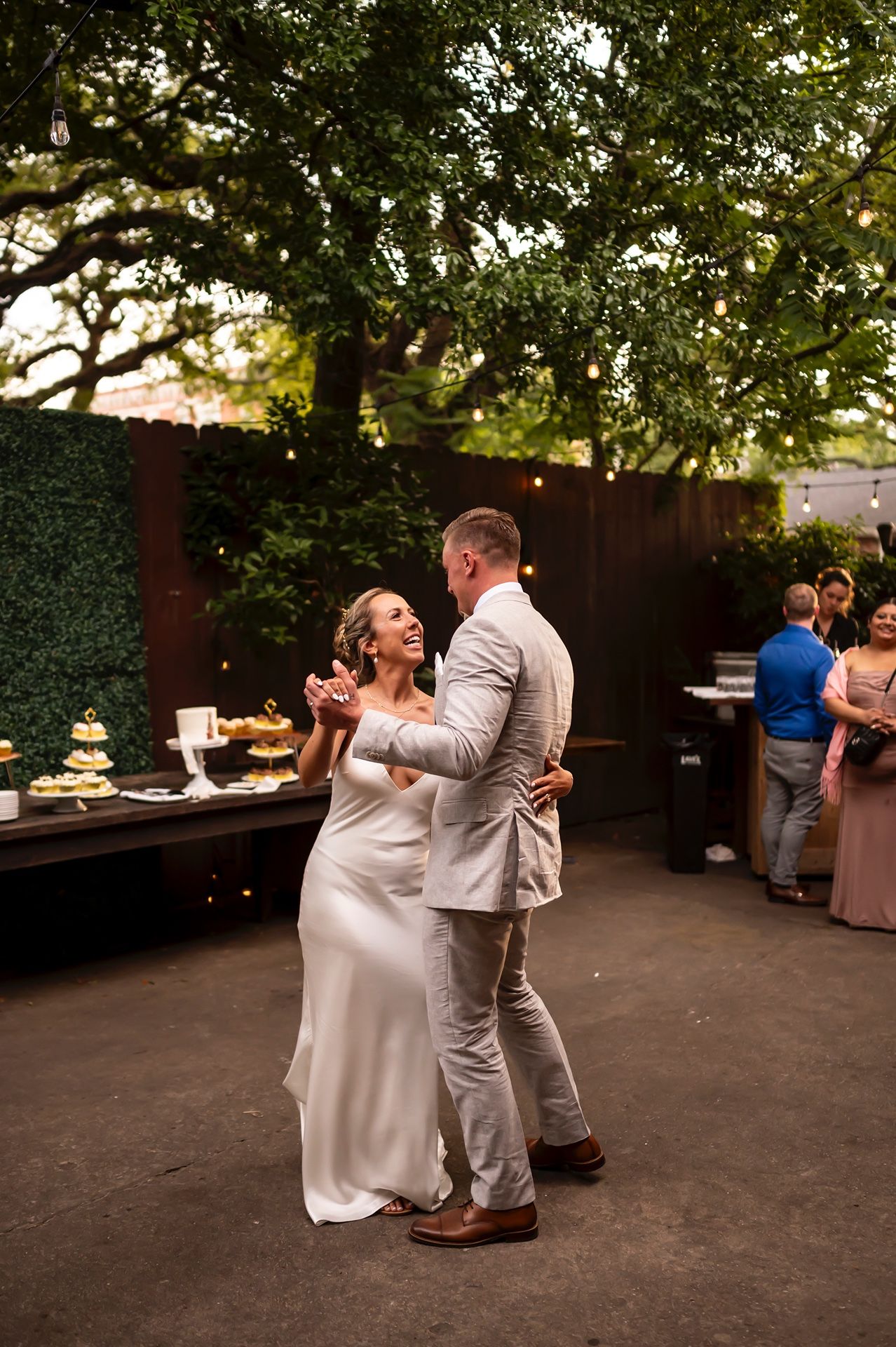 A couple shares a joyful dance moment at an outdoor wedding reception with trees and a dessert table in the background.