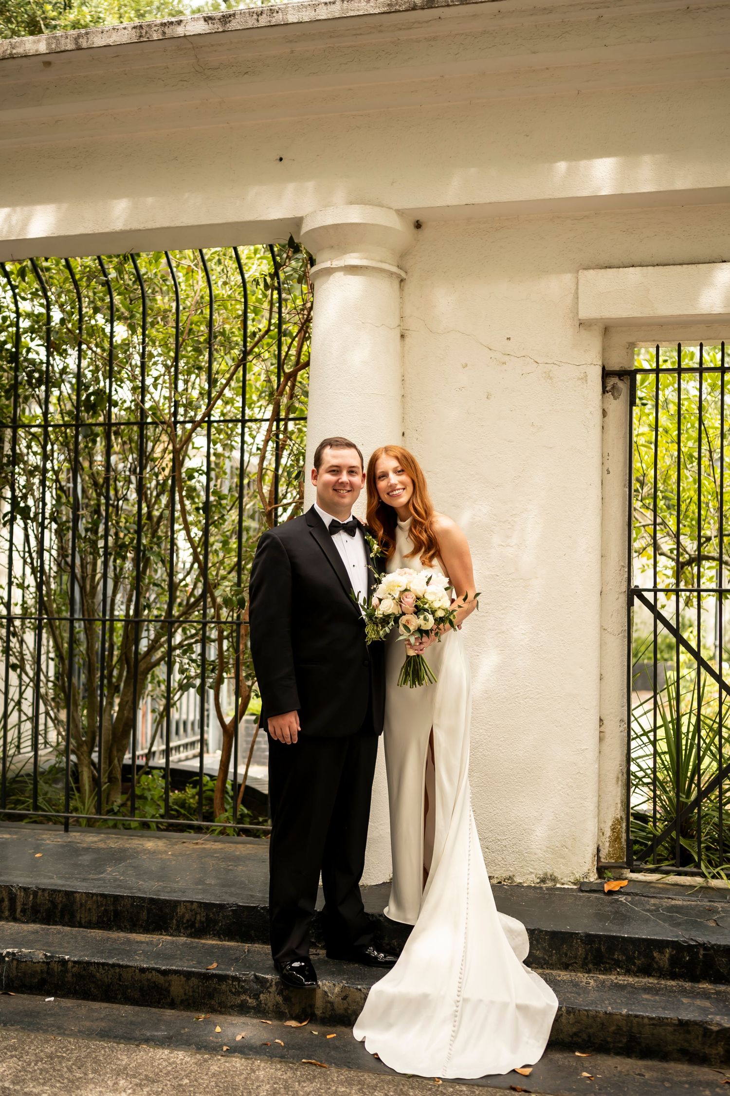 Couple poses for wedding photos in an elegant outdoor walkway with white stone pillars and lush greenery in the background.