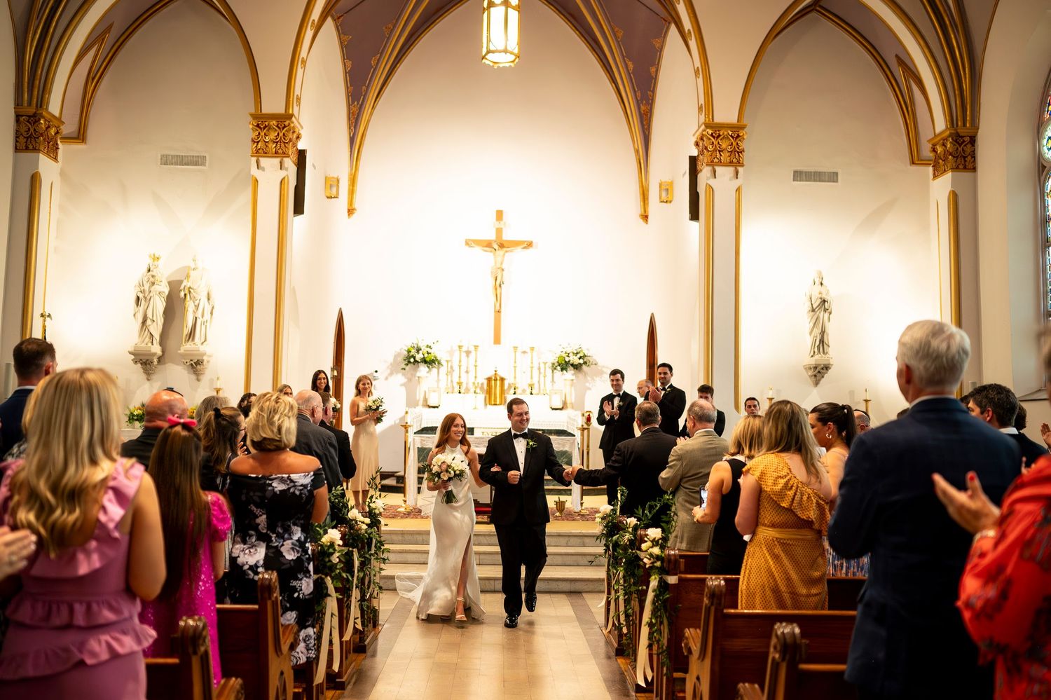 Wedding guests stand in church pews during ceremony at historic chapel with vaulted arches and ornate altar.