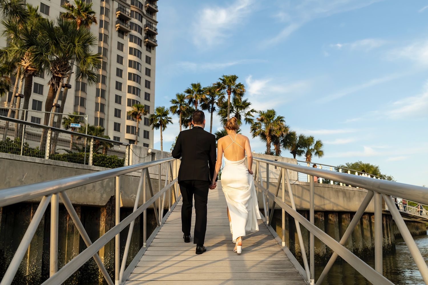 A couple walks along a wooden boardwalk lined with tall palm trees against a bright blue sky.