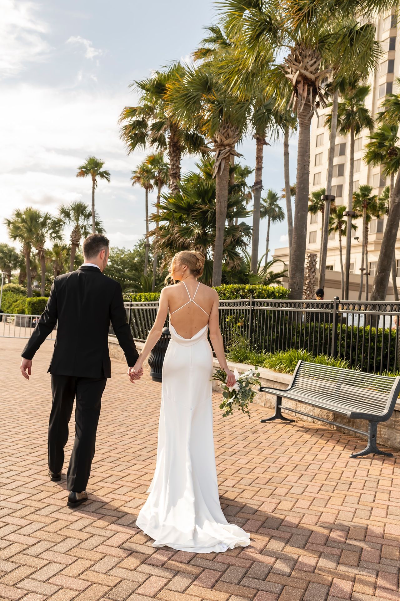 Four wedding photos show a couple exiting their venue along a scenic palm-lined brick path on their special day.