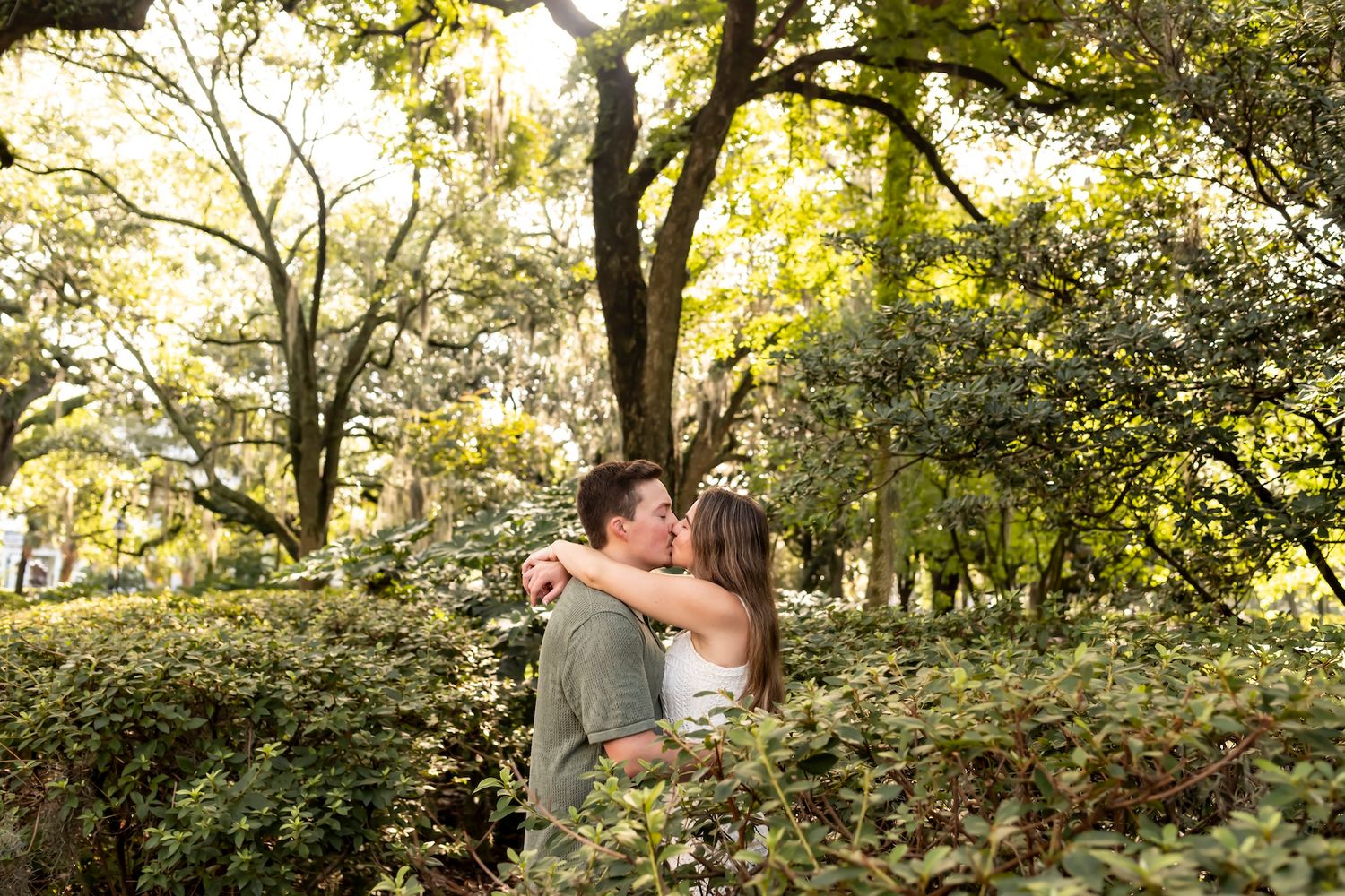A couple embraces in a sun-dappled garden setting surrounded by lush greenery and trees.