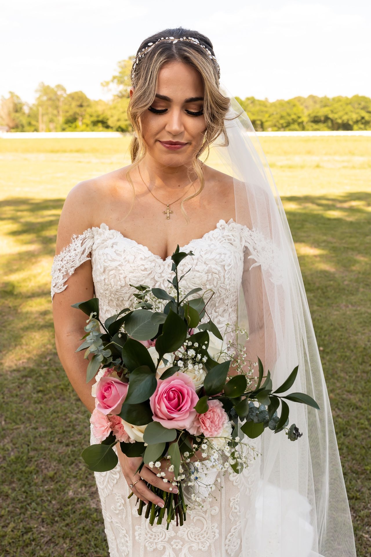 Beautiful lace wedding dress with off-shoulder detail and pink rose bouquet captured in outdoor garden setting.