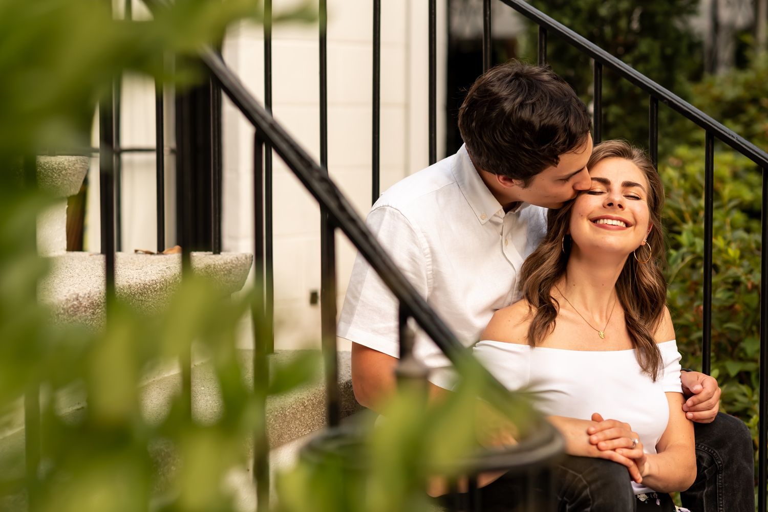 A romantic couple embraces while sitting on outdoor stairs surrounded by greenery in warm sunlight.