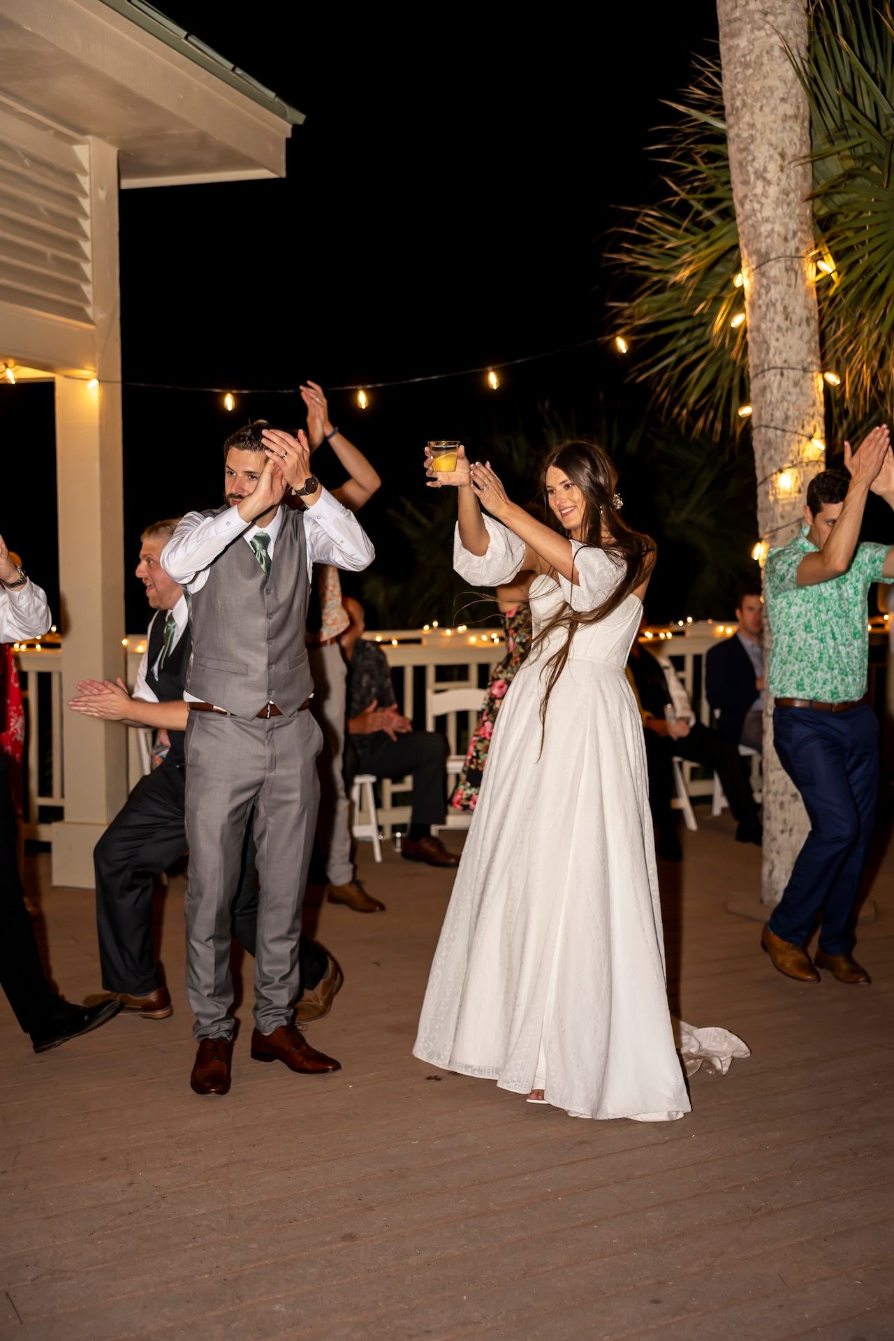 Wedding party dances at nighttime outdoor reception with string lights and palm trees visible on the patio.