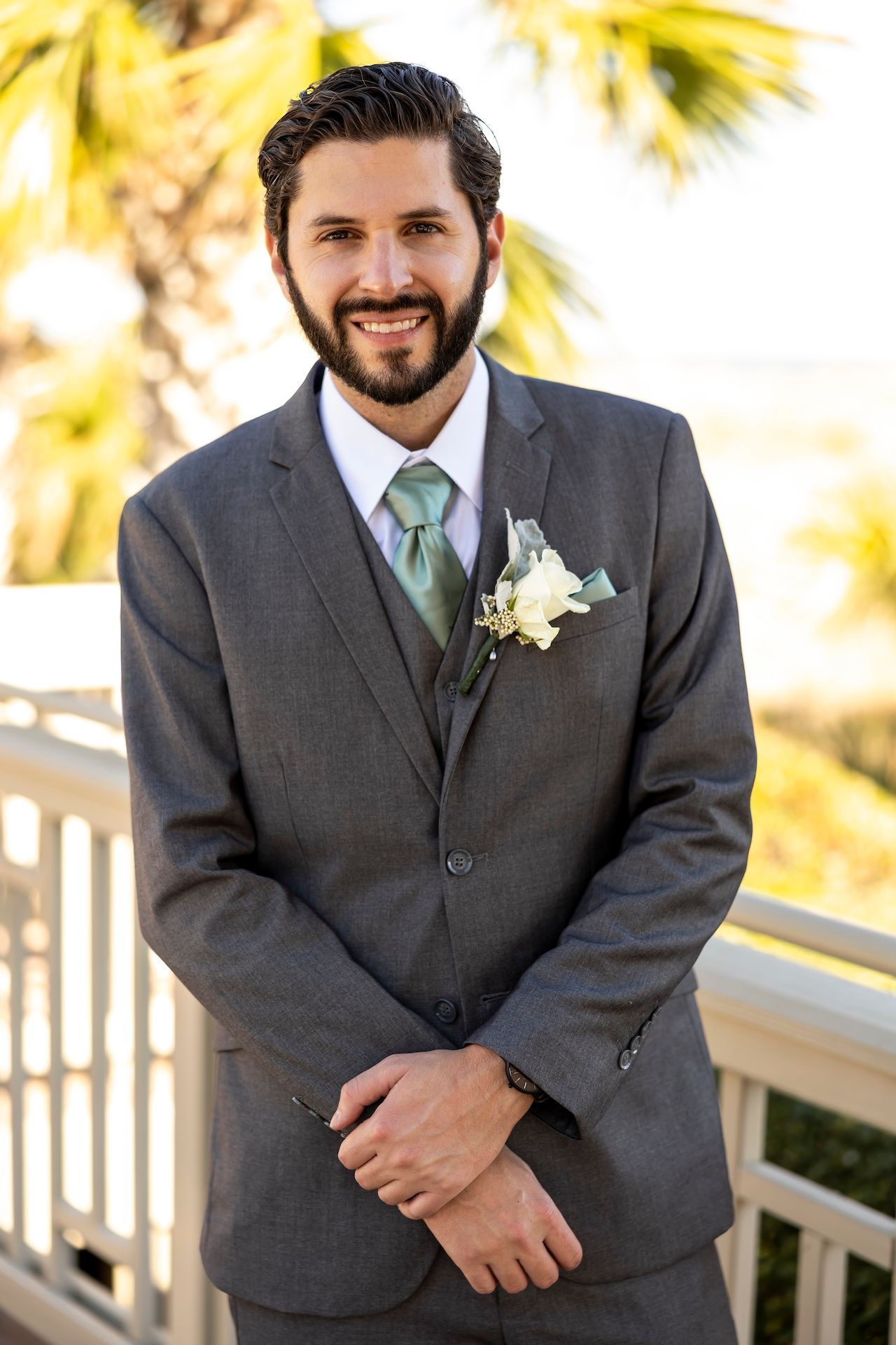 A person in a charcoal grey suit with green tie and white boutonniere smiles outdoors with palm trees in background.