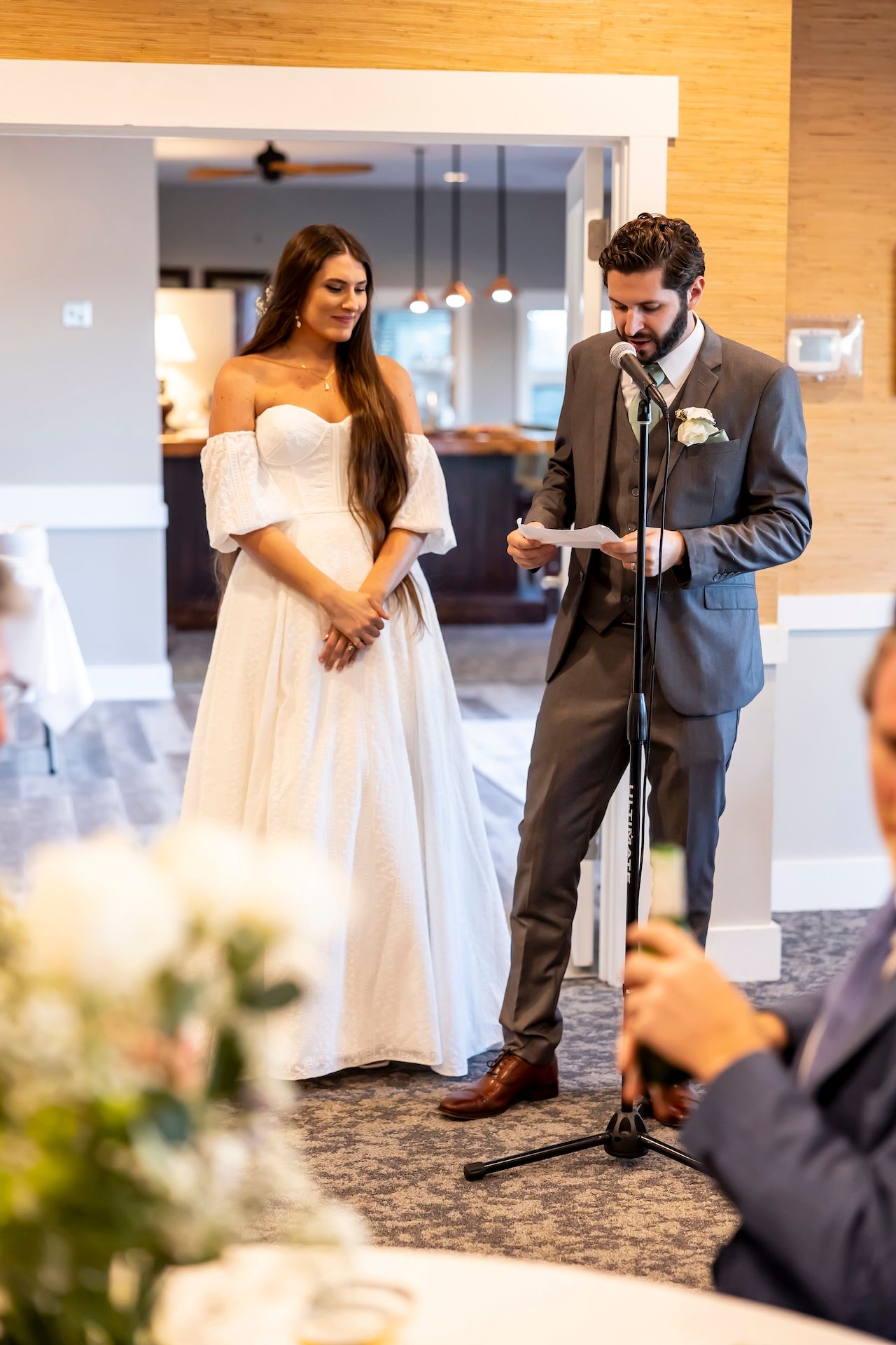 A bride in a flowing white gown stands beside her groom during their wedding ceremony as he reads from a paper.