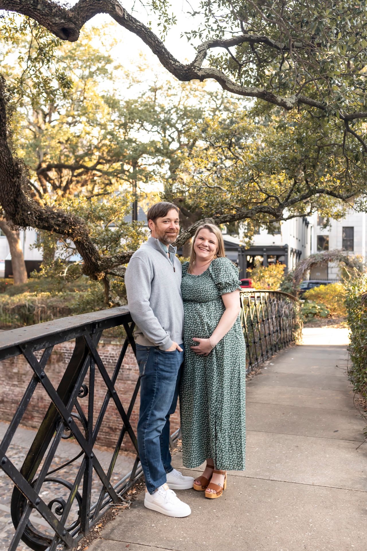 A couple stands together under a tree canopy on a garden pathway, with wrought iron railings and buildings in the background.