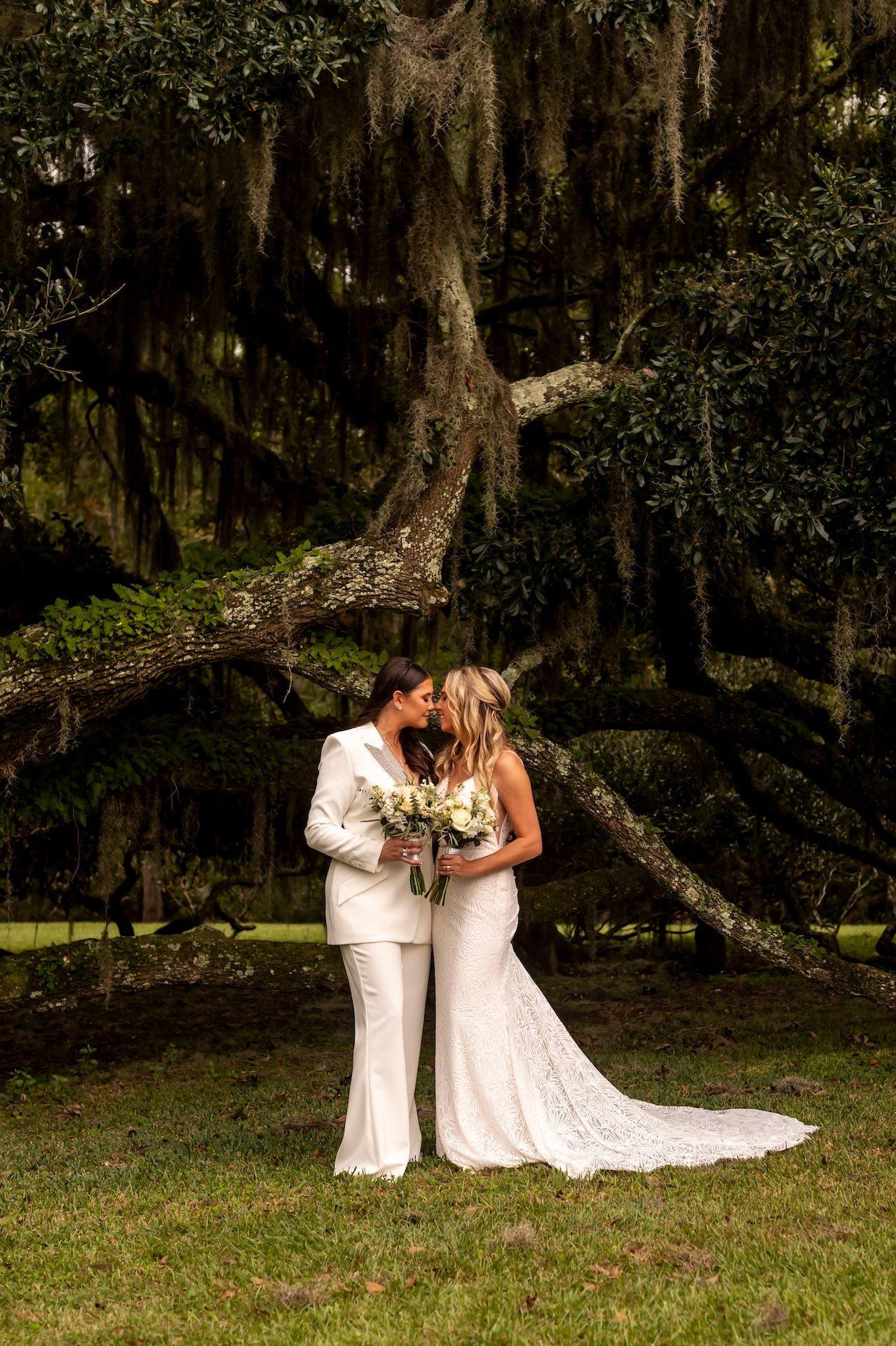 Two people in white attire share an intimate moment under a moss-draped oak tree in a romantic outdoor setting.